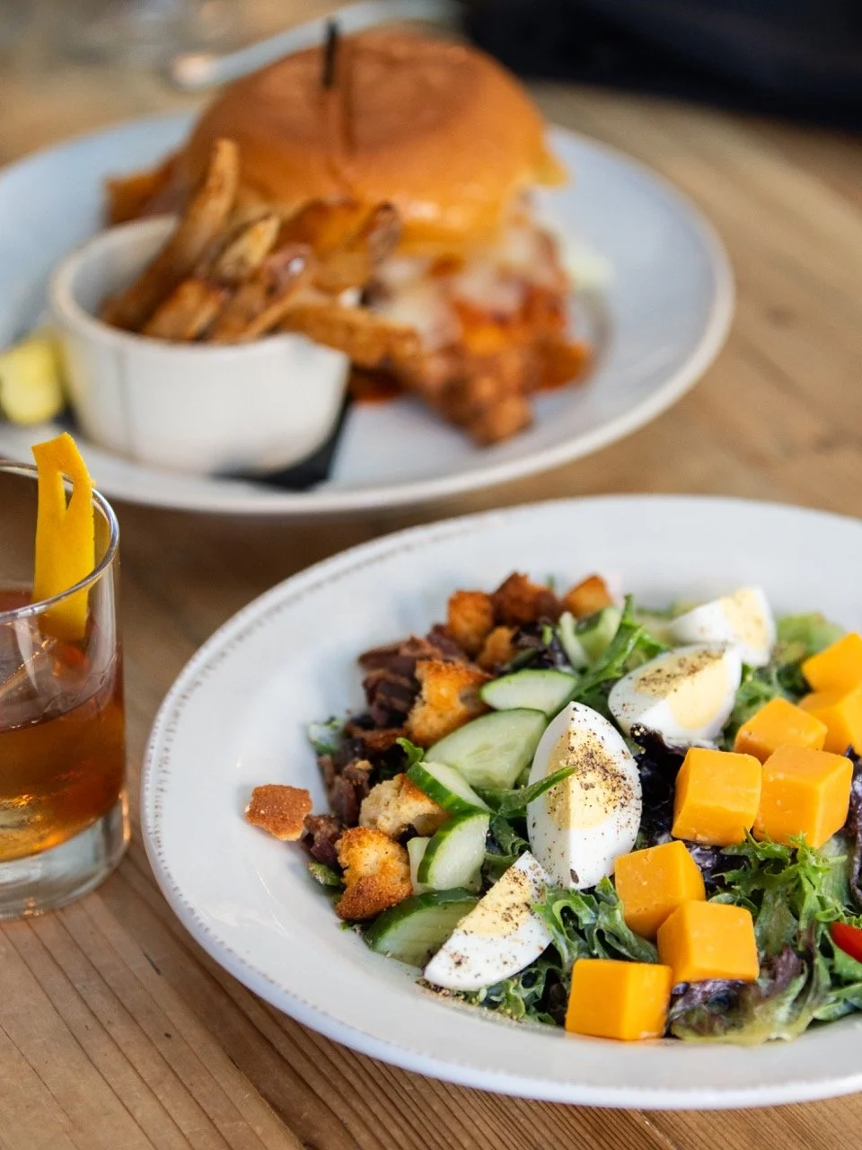 A plate of salad with cucumber, boiled eggs, cheddar cheese cubes, and mixed greens, with a glass of iced tea nearby, and another plate with a sandwich and fried chicken in the background.