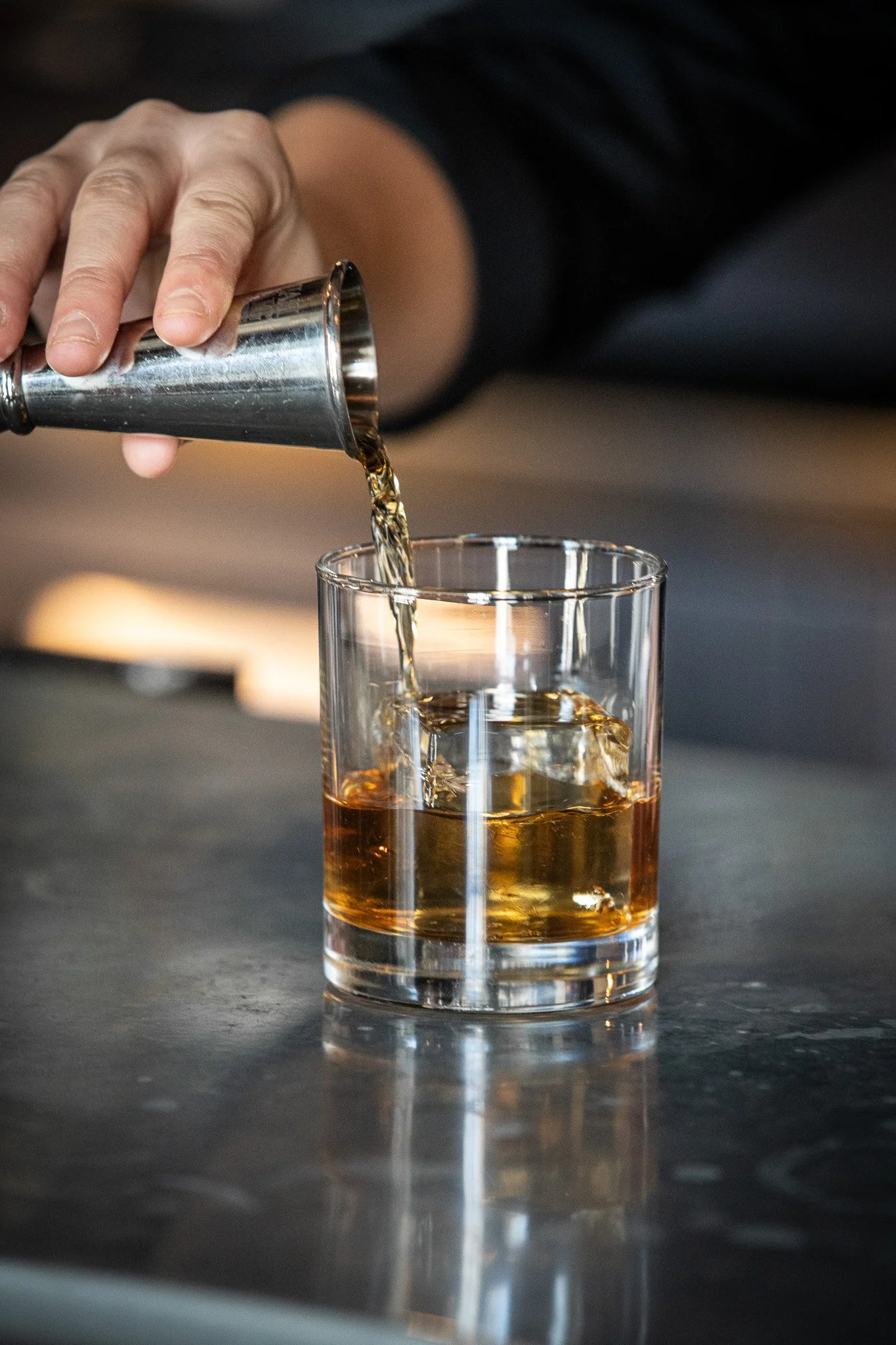 Hand pouring whiskey from a shaker into a glass with ice on a dark surface.
