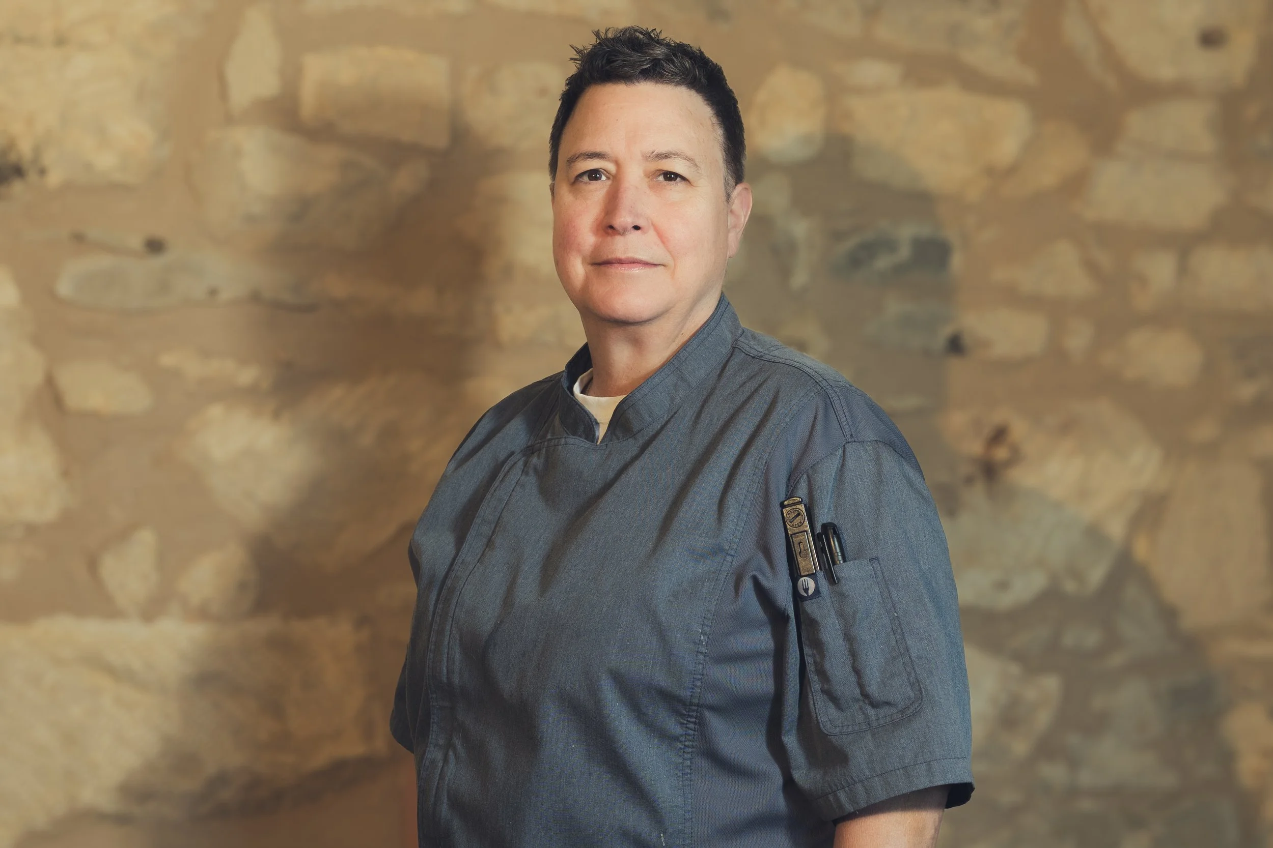 Portrait of a male chef in a gray chef's coat with a pen and thermometer in the sleeve pocket, standing against a stone wall background.