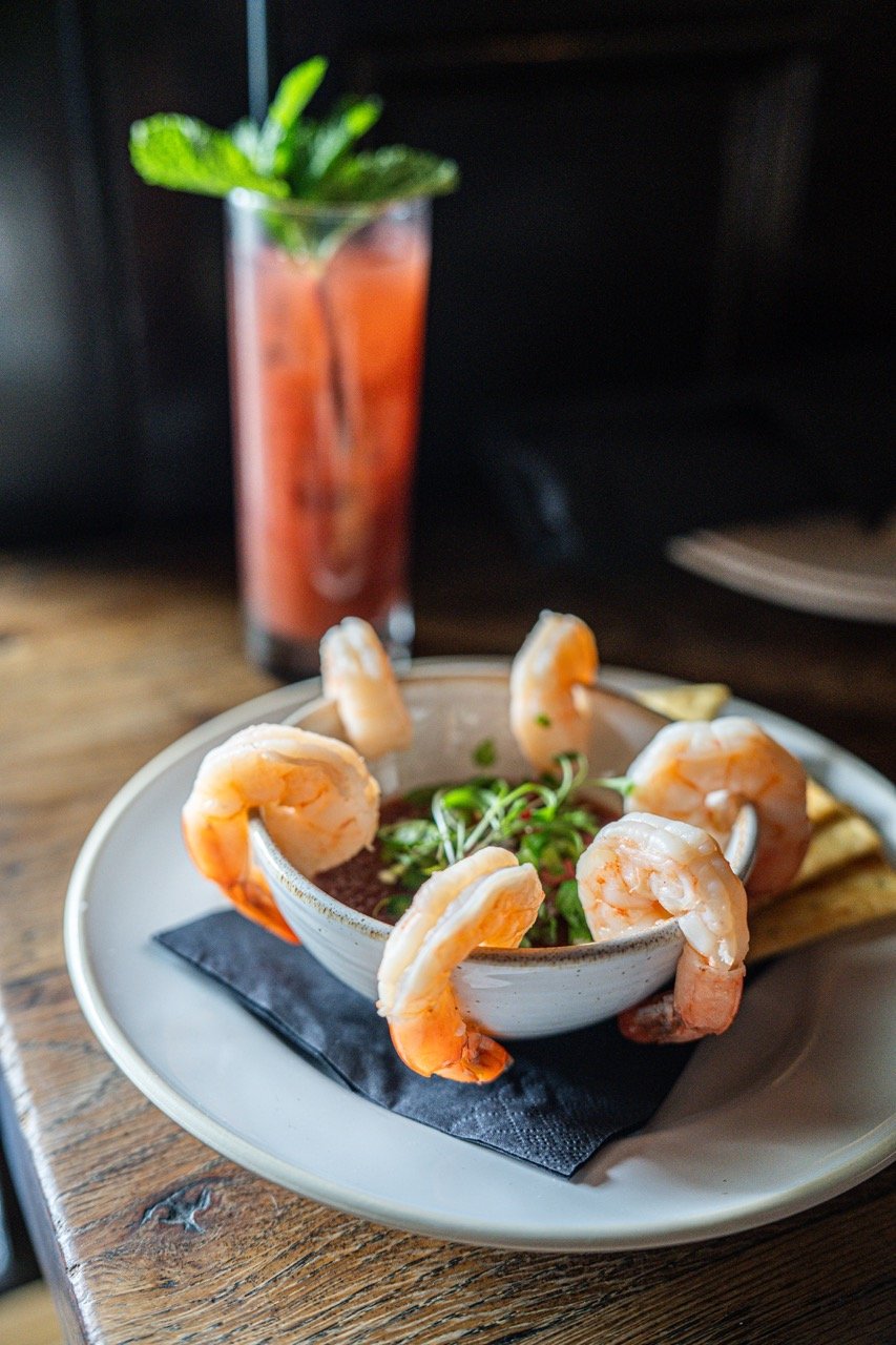 Shrimp cocktail with lemon wedges and a pink beverage with mint in a tall glass.