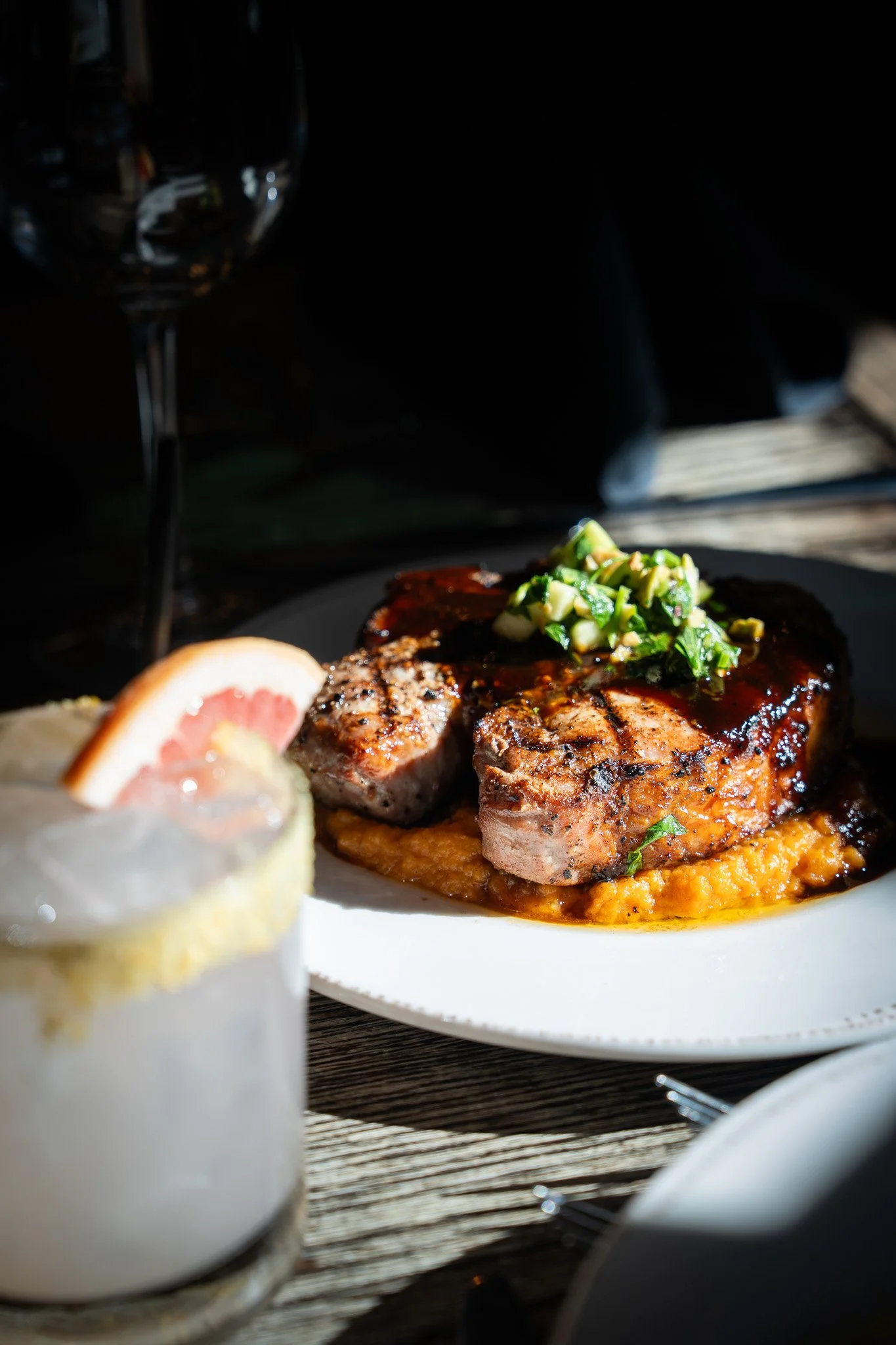 A plate with a grilled pork chop topped with chopped green onions served over mashed sweet potatoes. A cocktail garnished with a slice of grapefruit is also visible, with a wine glass and dark background.