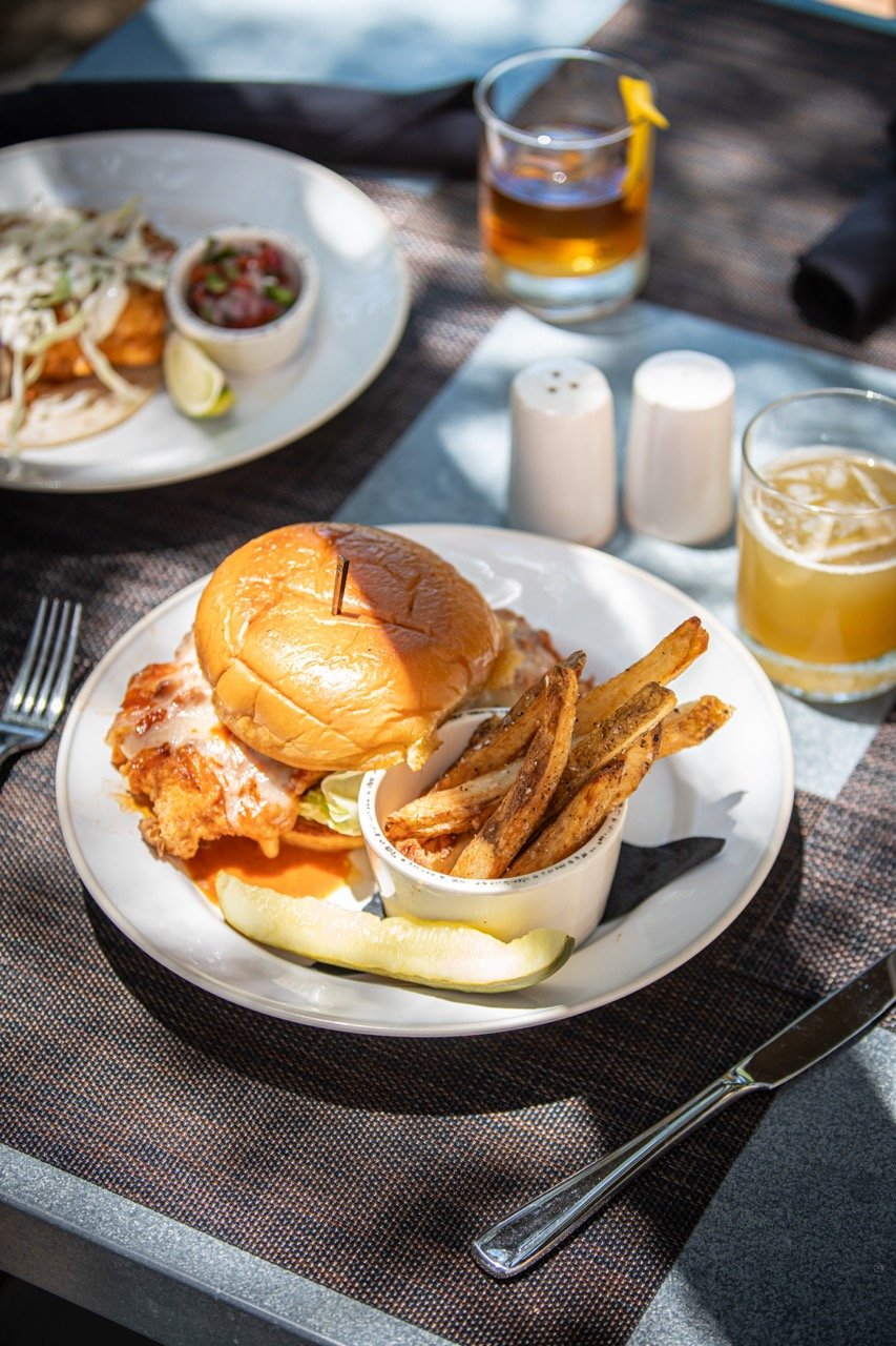 Fried chicken sandwich with lettuce and bun, a side of French fries in a white cup, a slice of lemon, and two drinks in glasses on a dark table