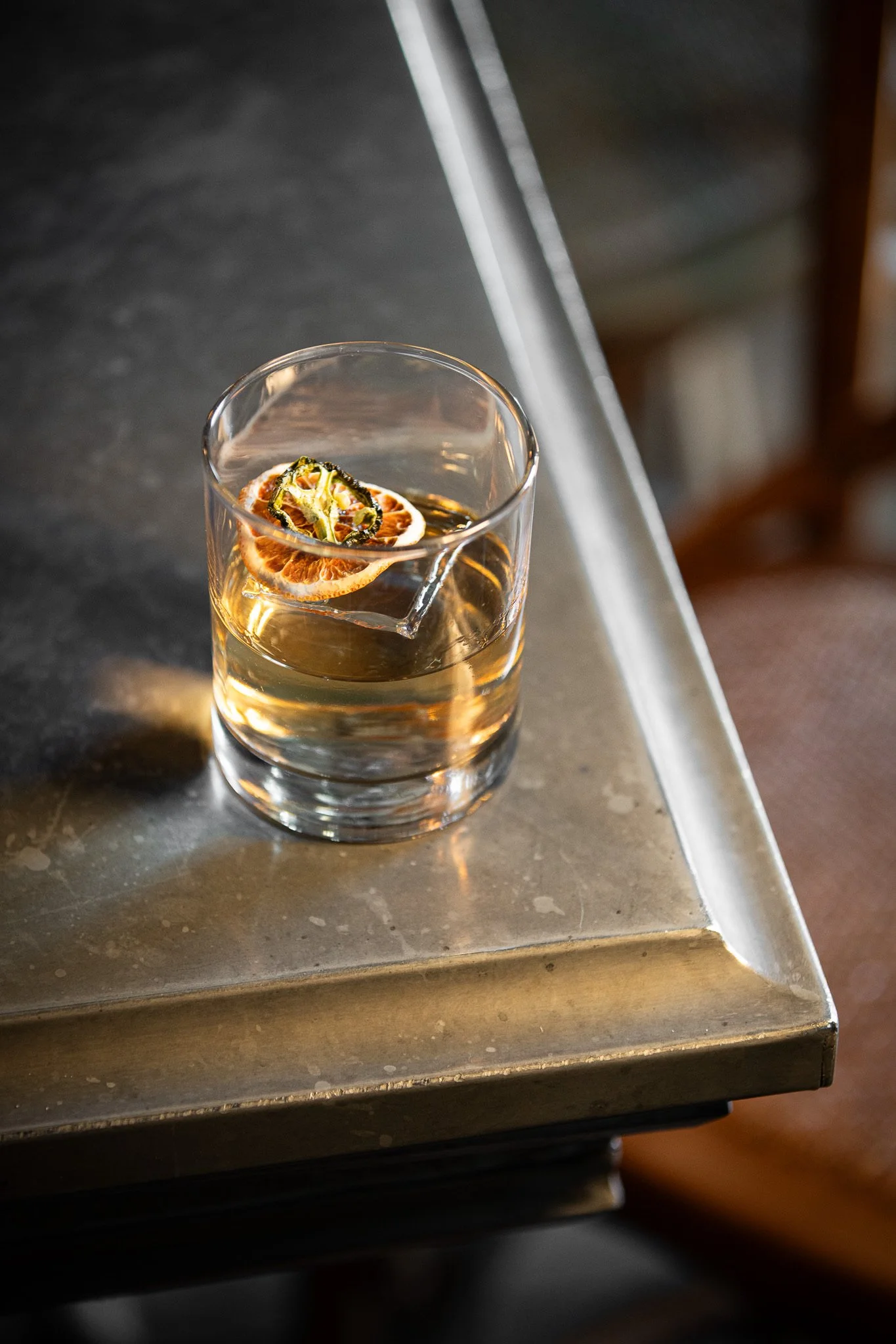 A clear glass of a light-colored beverage garnished with dried citrus slices and a green herb or vegetable, placed on a metal countertop.