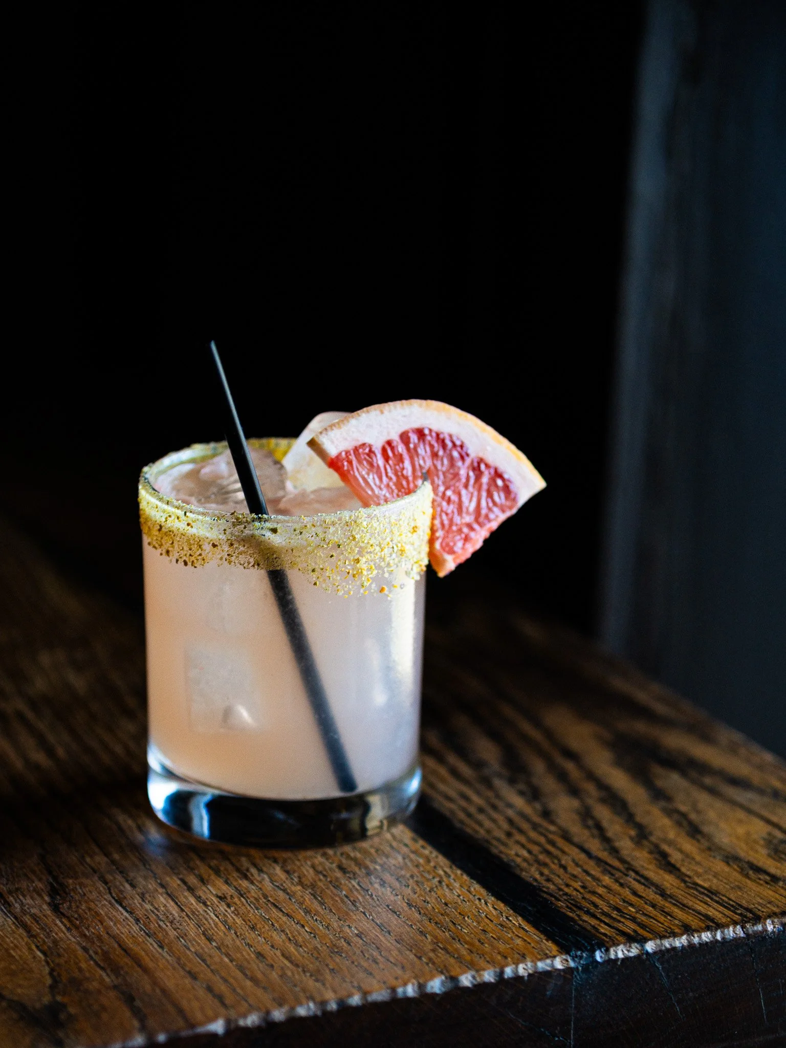 A glass of grapefruit cocktail with a slice of grapefruit garnish on the rim, pink drink with a black straw, on a wooden surface.