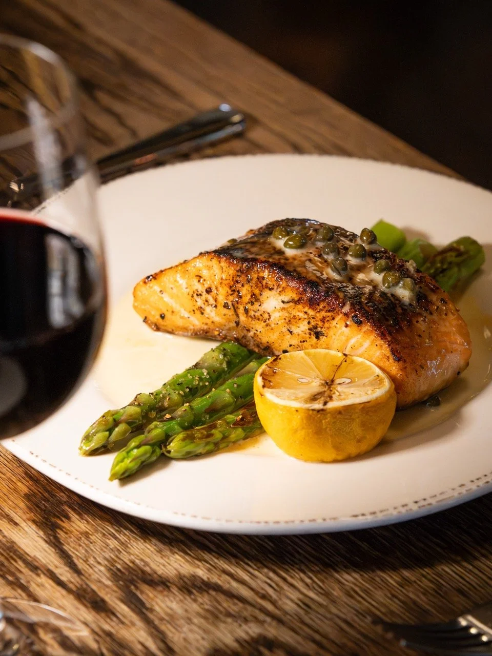 Grilled salmon fillet topped with capers, served with asparagus and a lemon half, with a glass of red wine on the side.