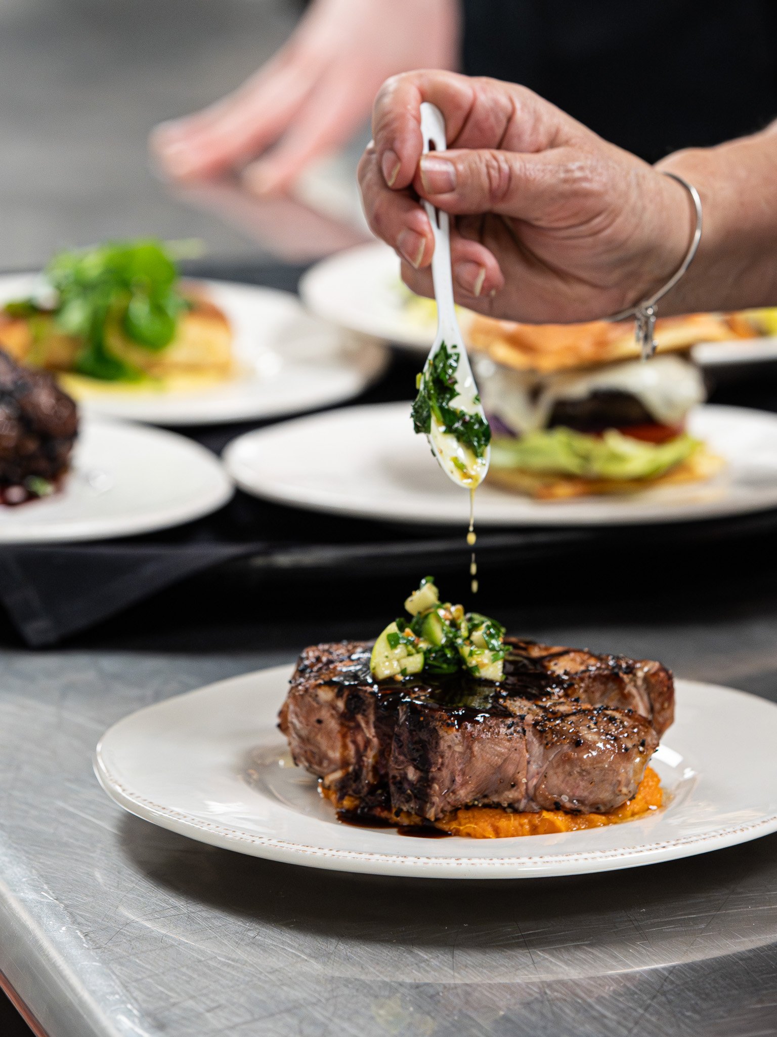 A chef's hand drizzling green herb sauce over a cooked steak topped with a small dice of vegetables on a white plate.
