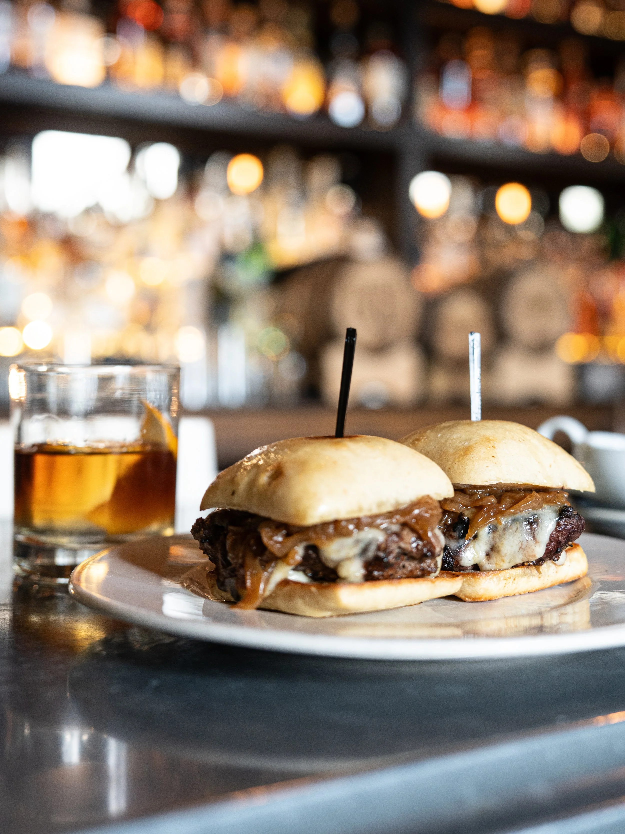 Close-up of two cheeseburgers with caramelized onions and melted cheese on a white plate, with a glass of iced tea and a blurred bar in the background.