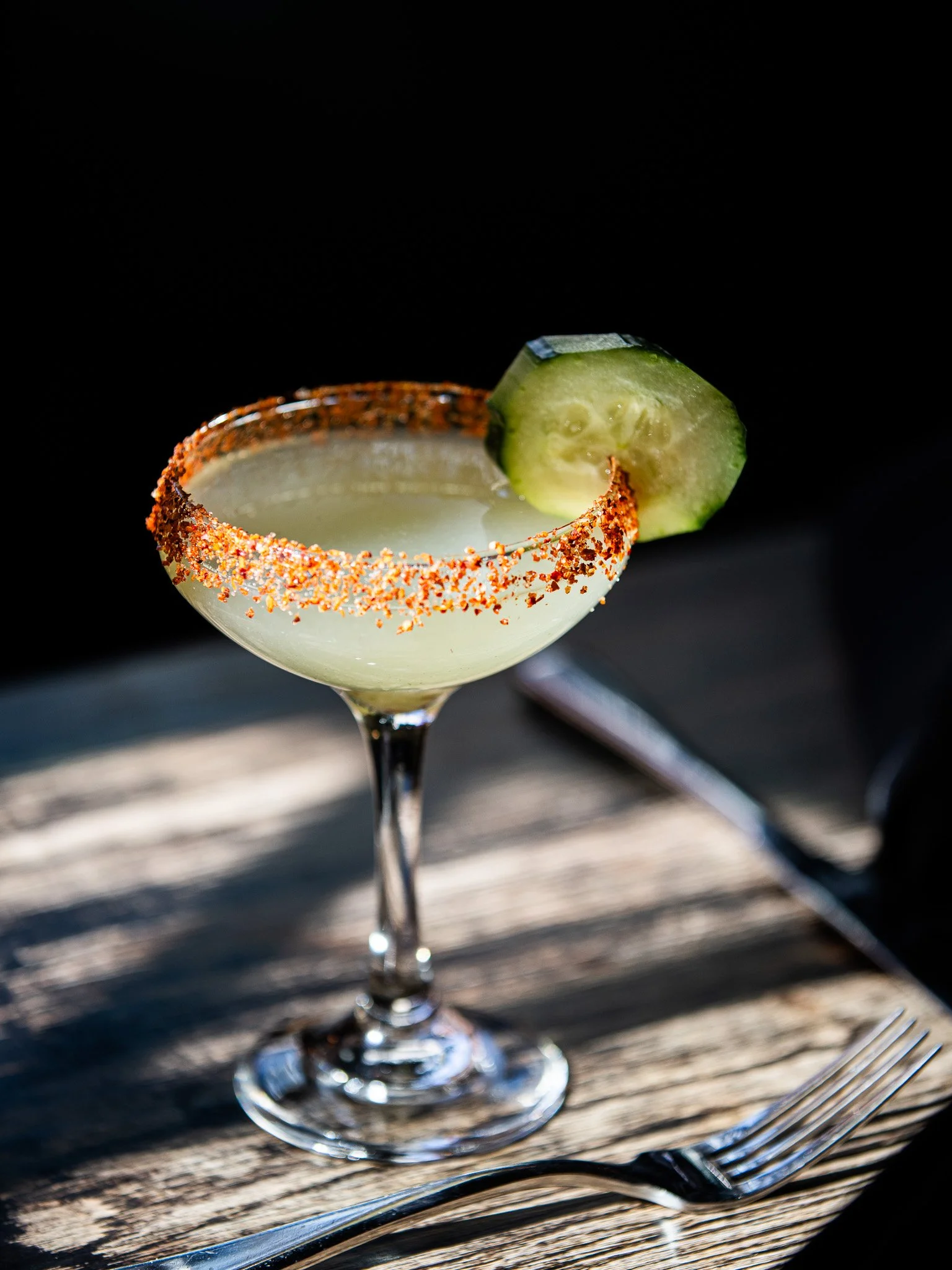A margarita cocktail in a classic glass with a salted rim and a cucumber slice garnish on the rim, placed on a wooden table.