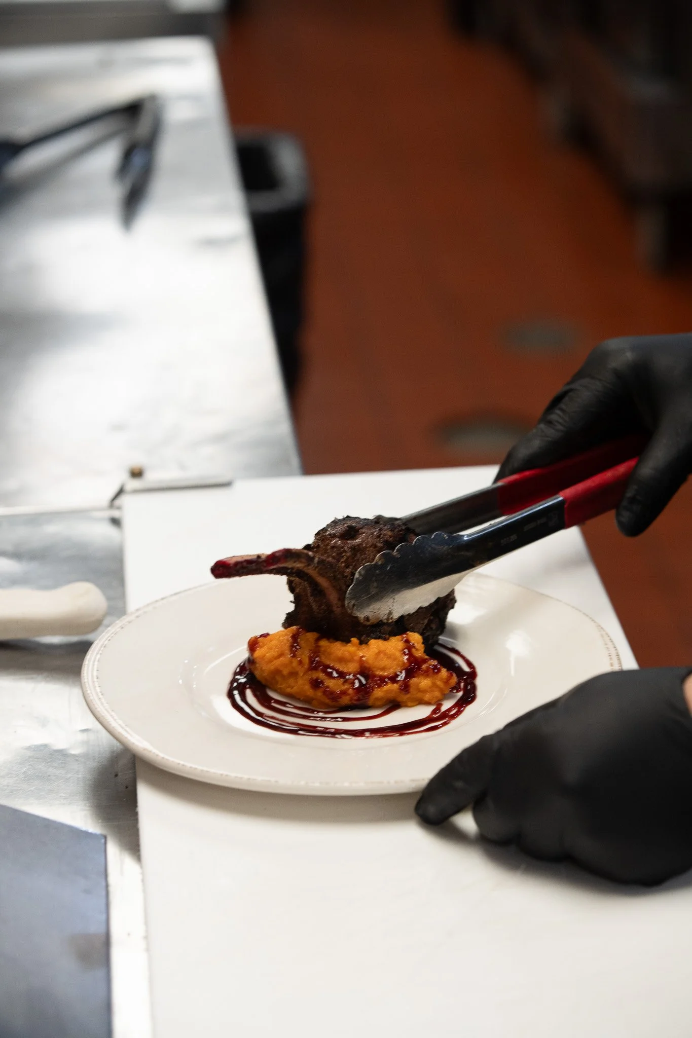 Chef wearing black gloves serving a grilled lamb chop on a bed of sweet potatoes with a drizzle of sauce on a white plate.