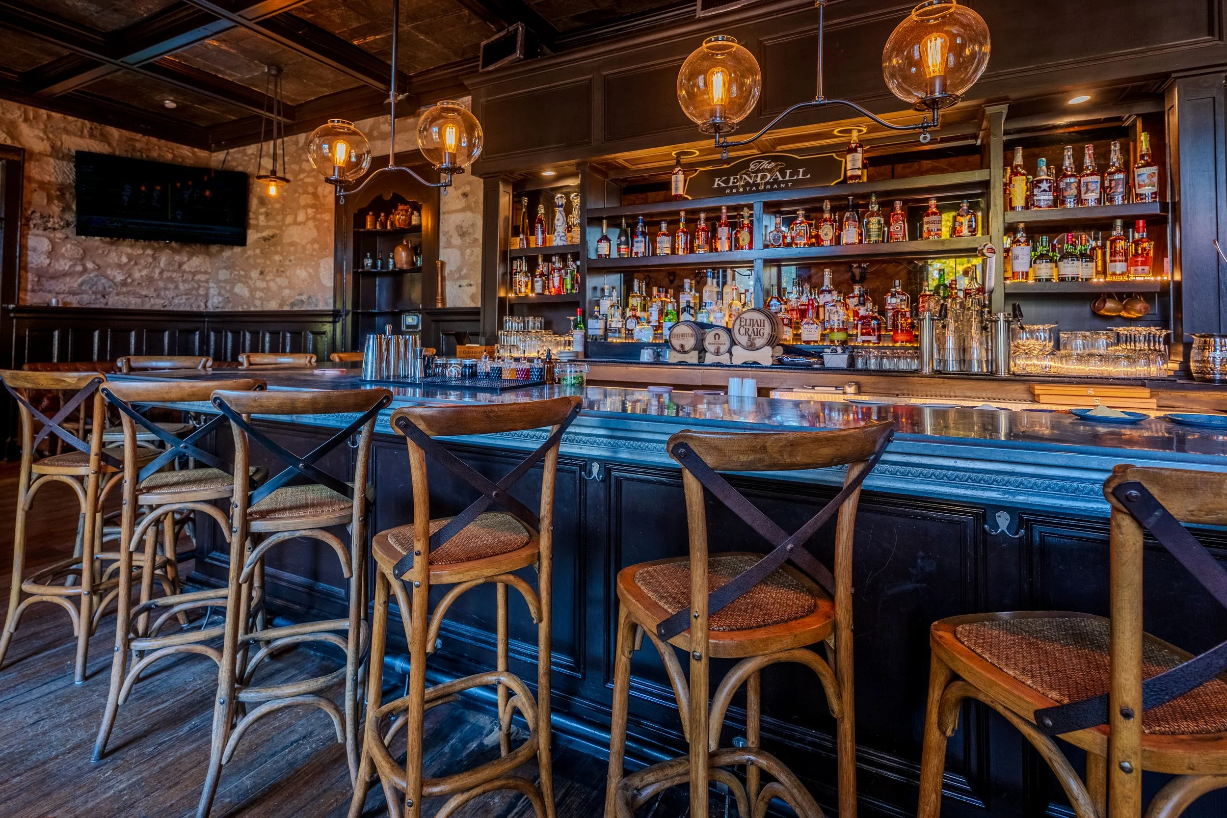 Interior of a cozy bar with a polished marble counter, wooden bar stools, and a dark wood back bar stocked with various bottles of liquor. Pendant lights with glass globes hang from the ceiling, and a stone wall is visible in the background.