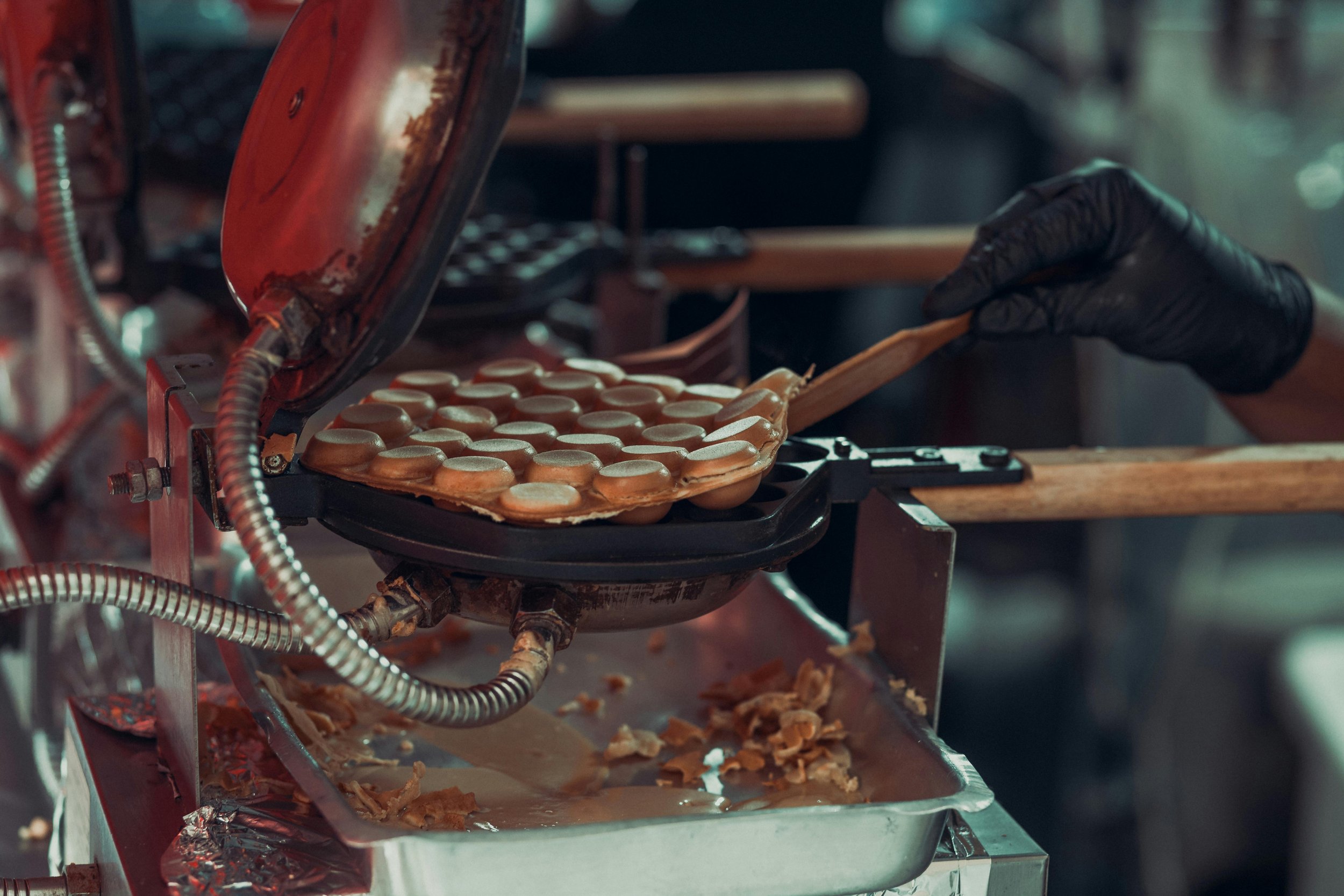 A person wearing black gloves is using a wooden spatula to flip small, round, light-colored food items, possibly eggs, on a black industrial cooking device in a kitchen. The device is releasing steam or smoke, and the surface has some debris and food scraps.
