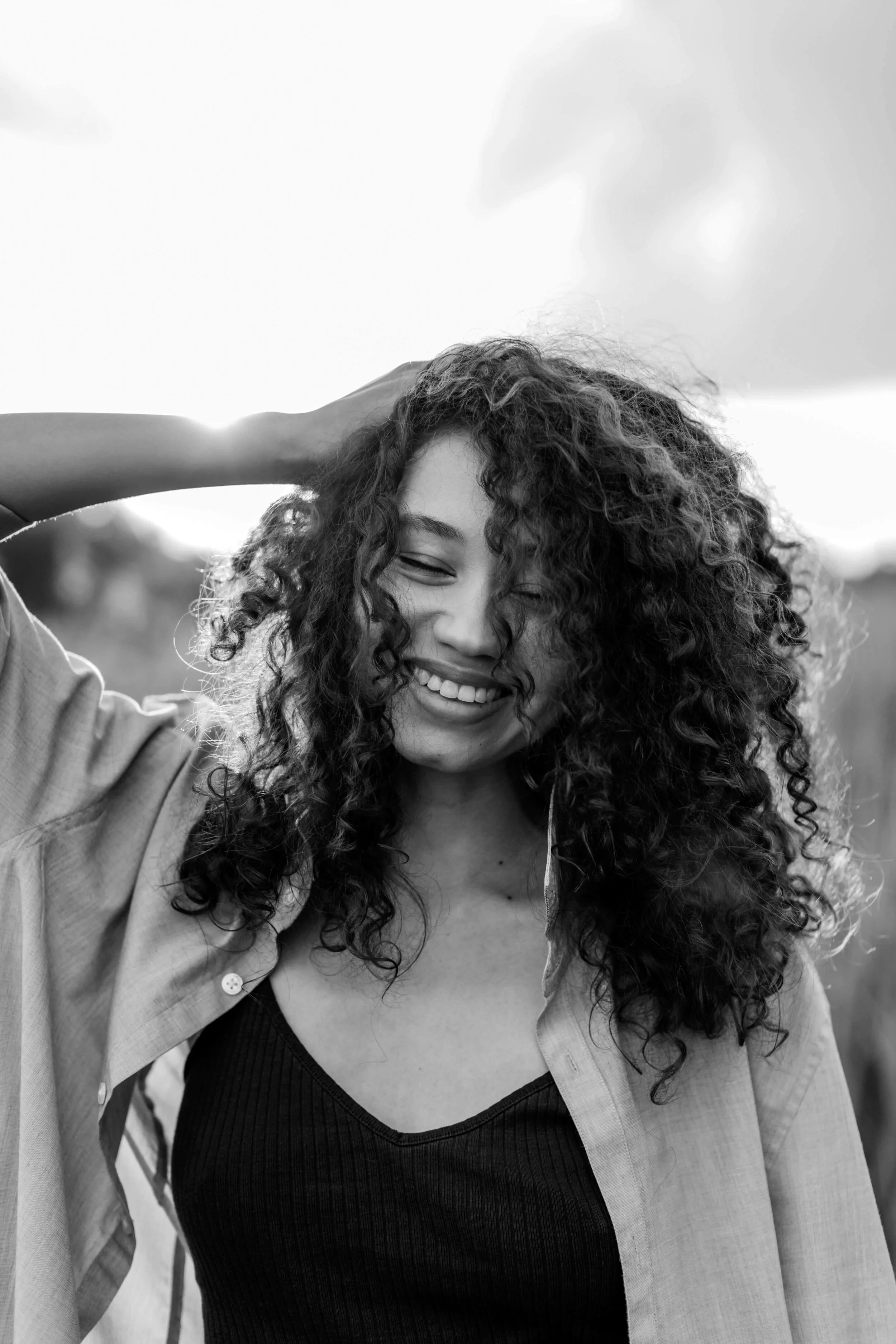 A smiling woman with curly hair, wearing a black top and light-colored shirt, standing outdoors.