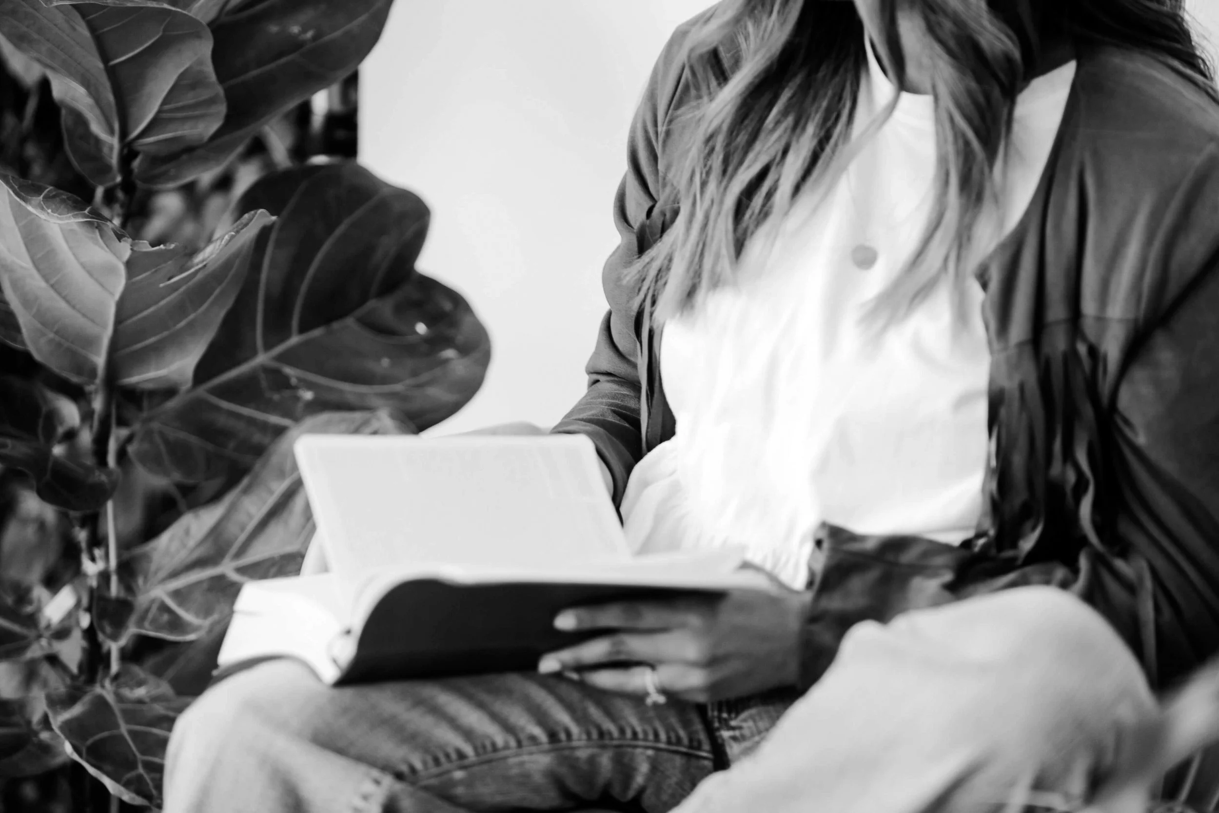 A person reading a book while sitting next to large leafy plants.