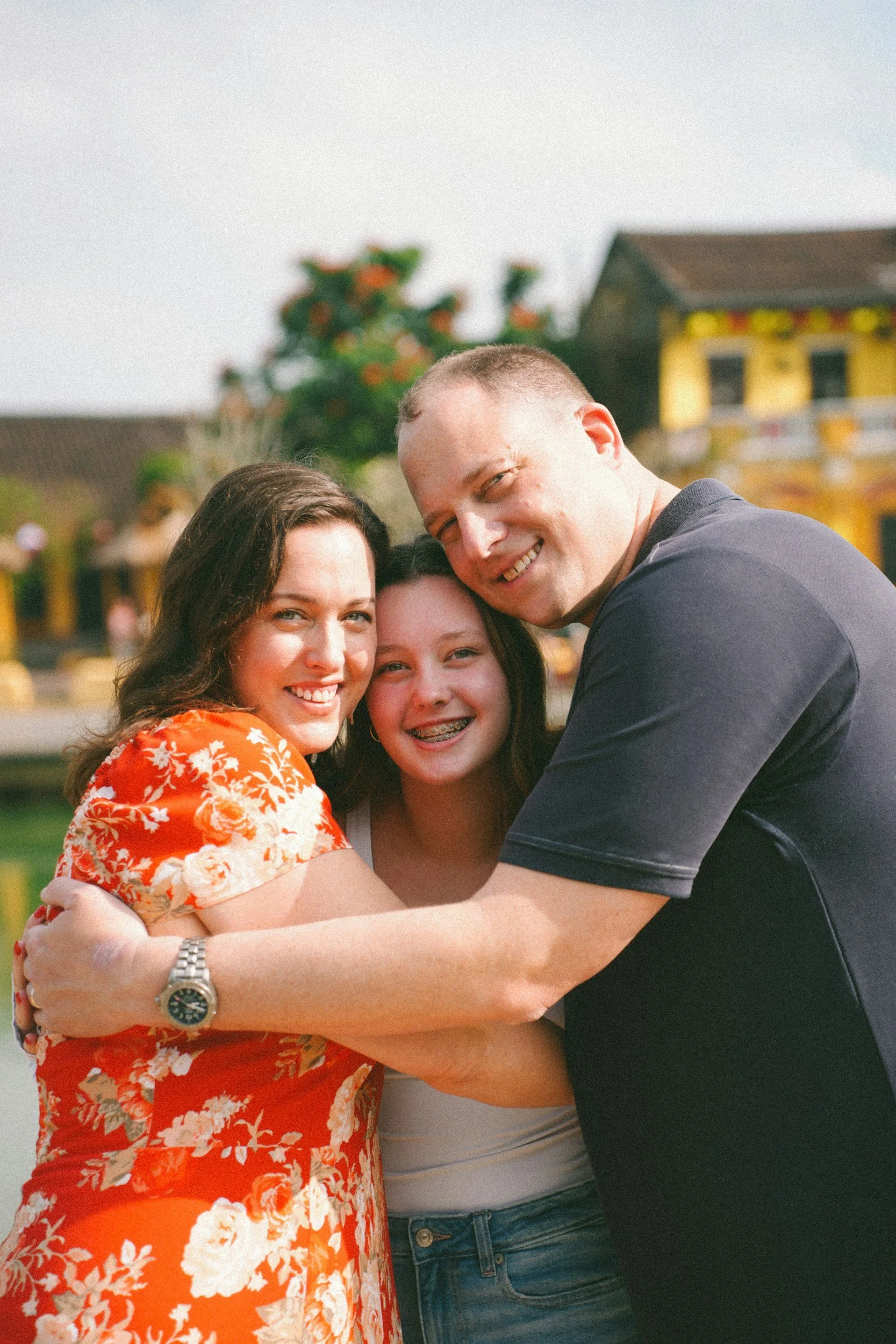 A family of three hugging outdoors, smiling at the camera, with colorful houses in the background.