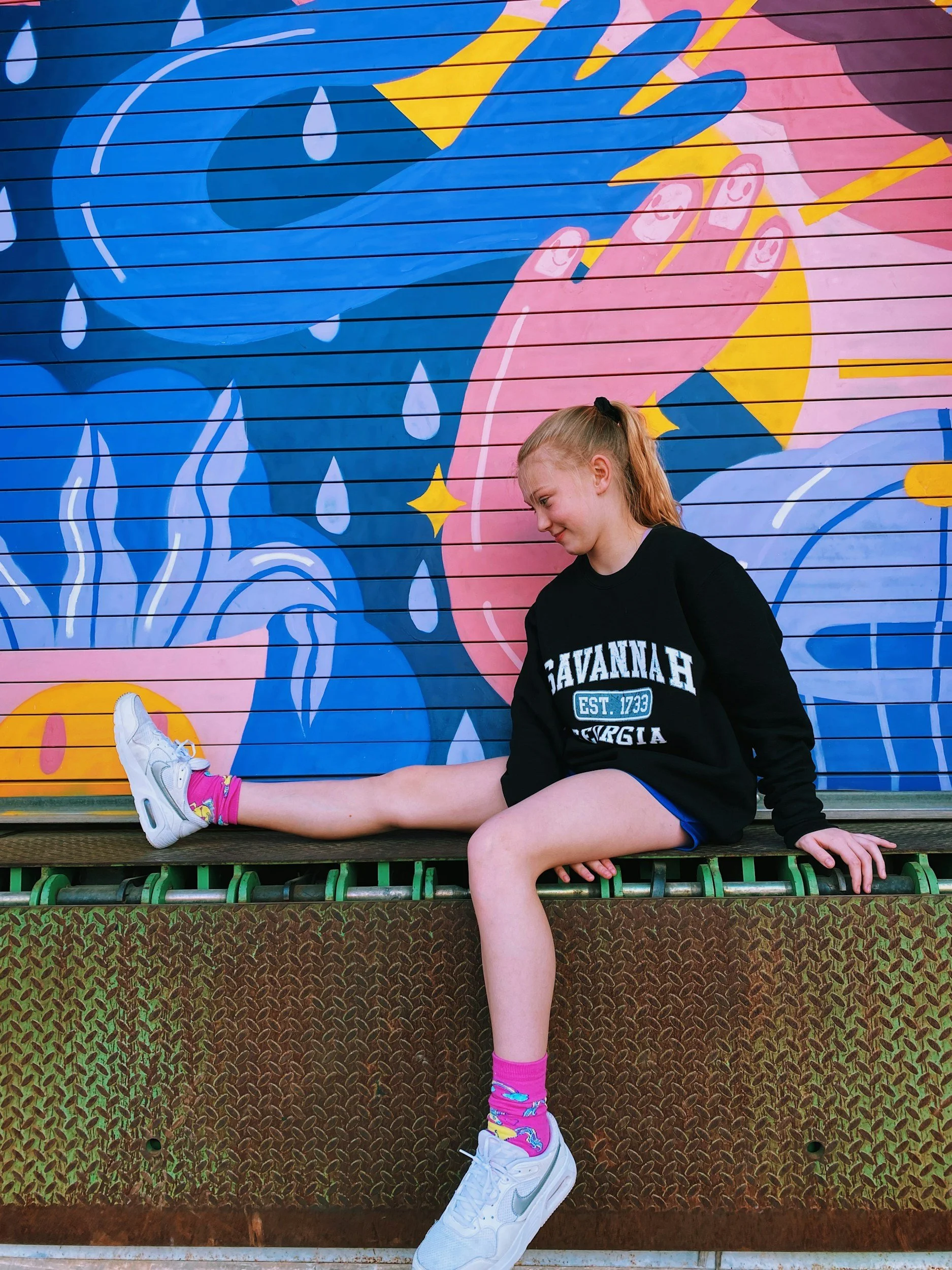 A young girl sitting with one leg extended and the other bent, posing in front of a colorful mural of a woman crying with her hand on her face and a rainy scene.