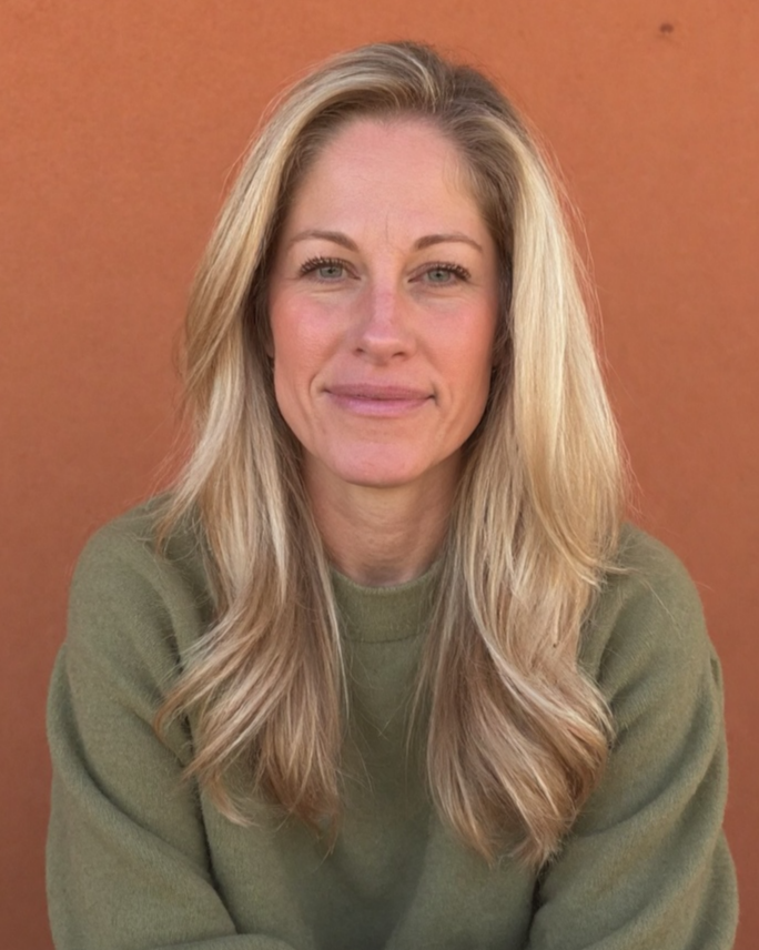 A woman with long blonde hair, wearing an olive green top, smiling softly, against an orange wall background.