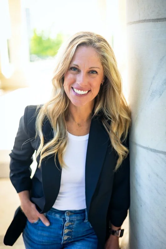 A smiling blonde woman in a black blazer and white shirt leans against a white wall.