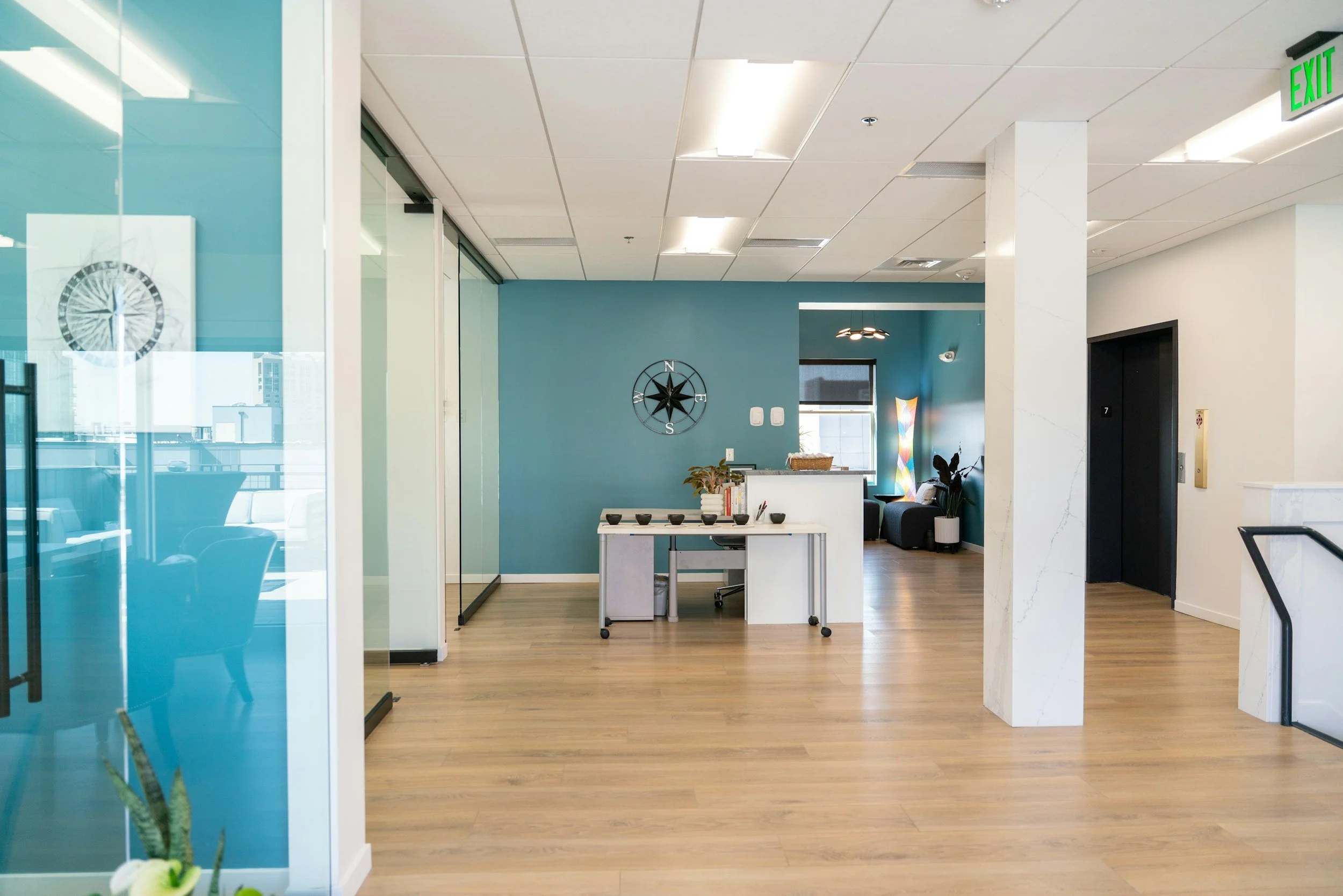A modern office lobby with blue accent walls, wooden floors, a small reception desk, and decorative elements like plants and artwork.