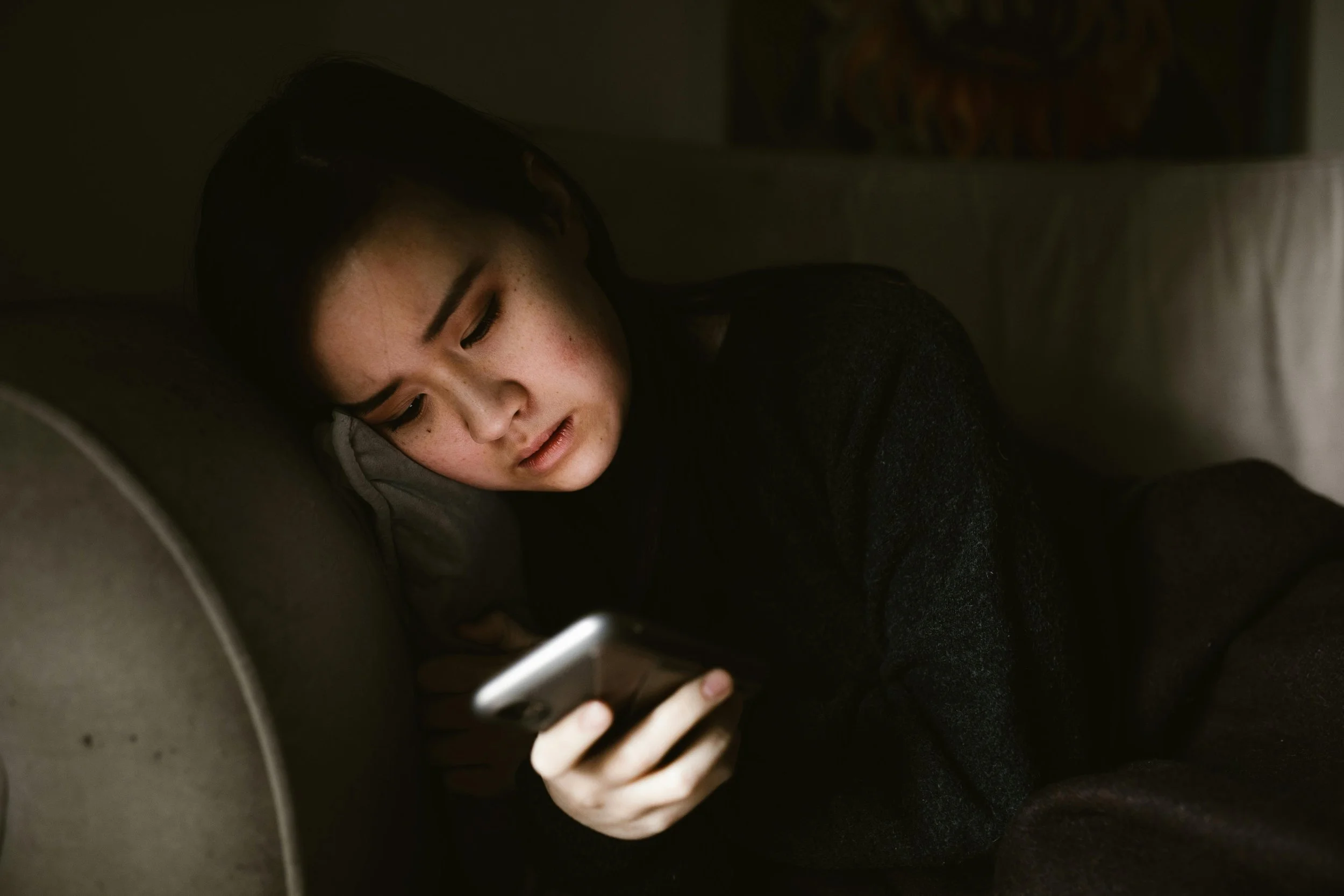 A woman sitting in a dimly lit room, looking reflective while holding a phone, representing the internal emotional intensity and relational fragility often felt during the luteal phase of the menstrual cycle.