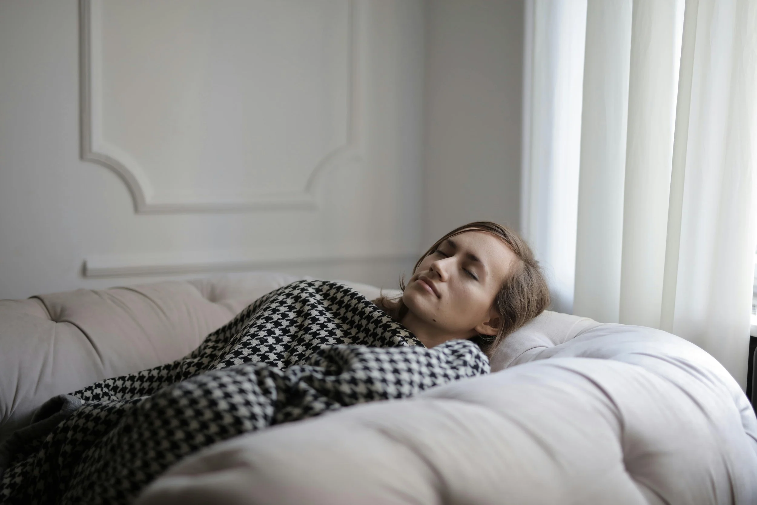 A woman lying down on a sofa with her eyes closed, wrapped in a blanket, illustrating the emotional exhaustion and somatic collapse that often occurs when transitioning from a high-pressure work day to home life.