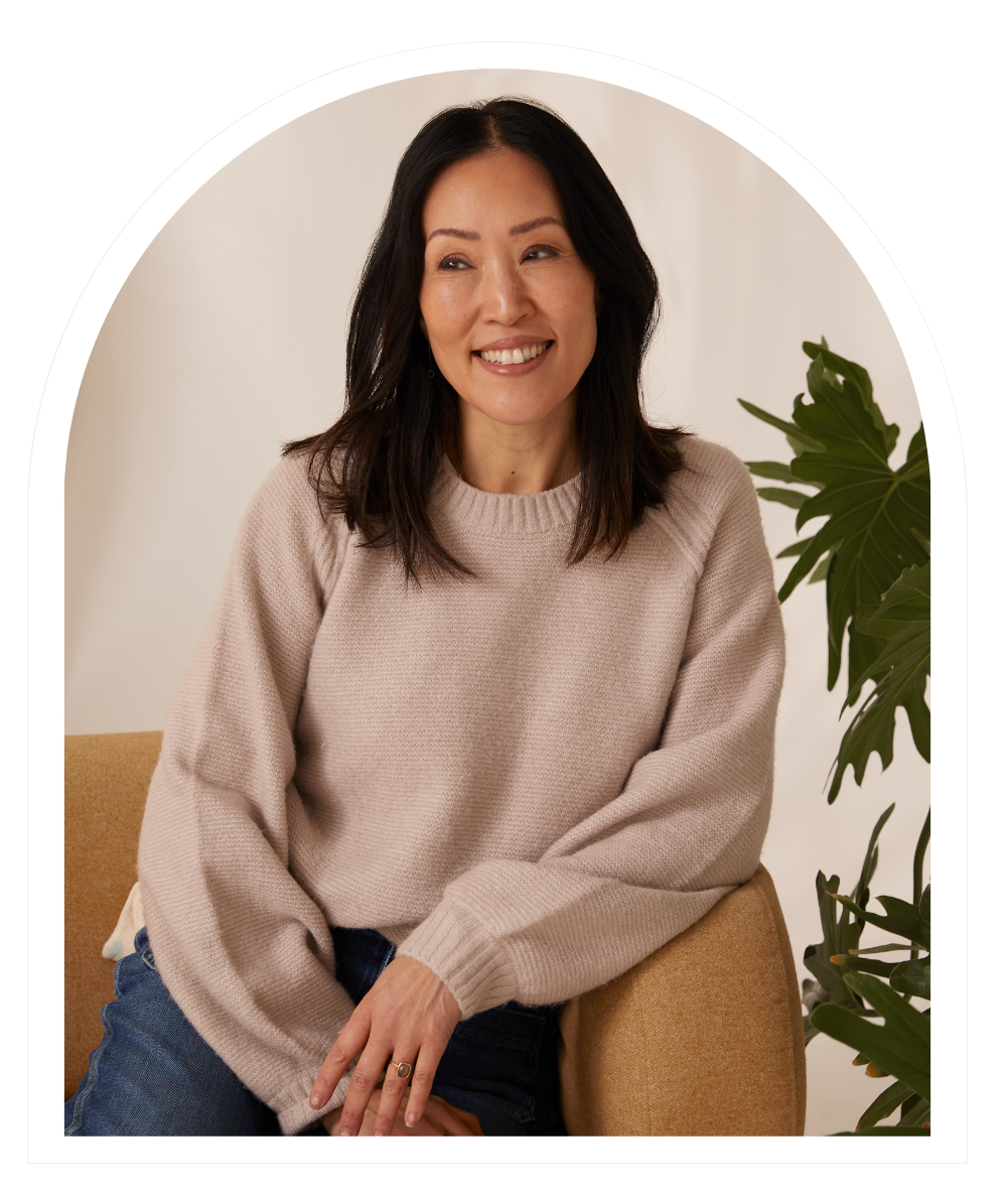 A woman with shoulder-length dark hair, wearing a beige sweater, sitting on an orange chair next to a green houseplant, smiling at the camera.