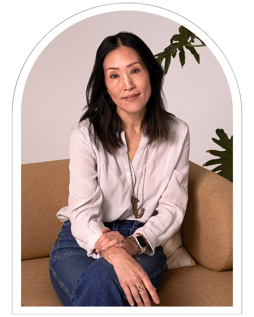 Catherine SoYoung, A woman with shoulder-length dark hair, wearing a cream blouse, blue jeans, and jewelry, sitting on a beige sofa with a large green plant in the background.