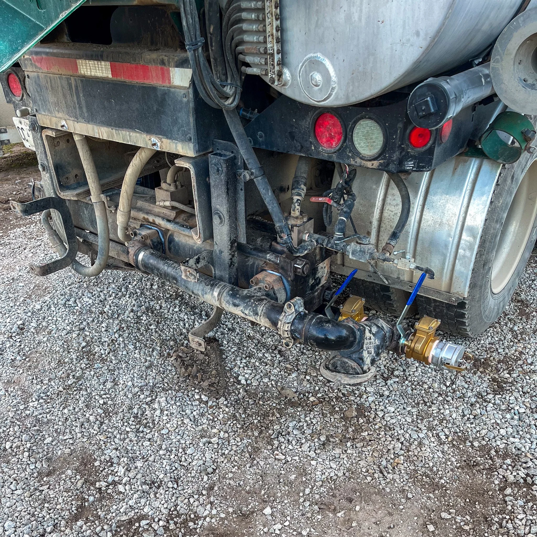 Close-up of the rear section of a utility truck showing hydraulic and fuel system components, hoses, pipes, and wiring on the gravel ground.
