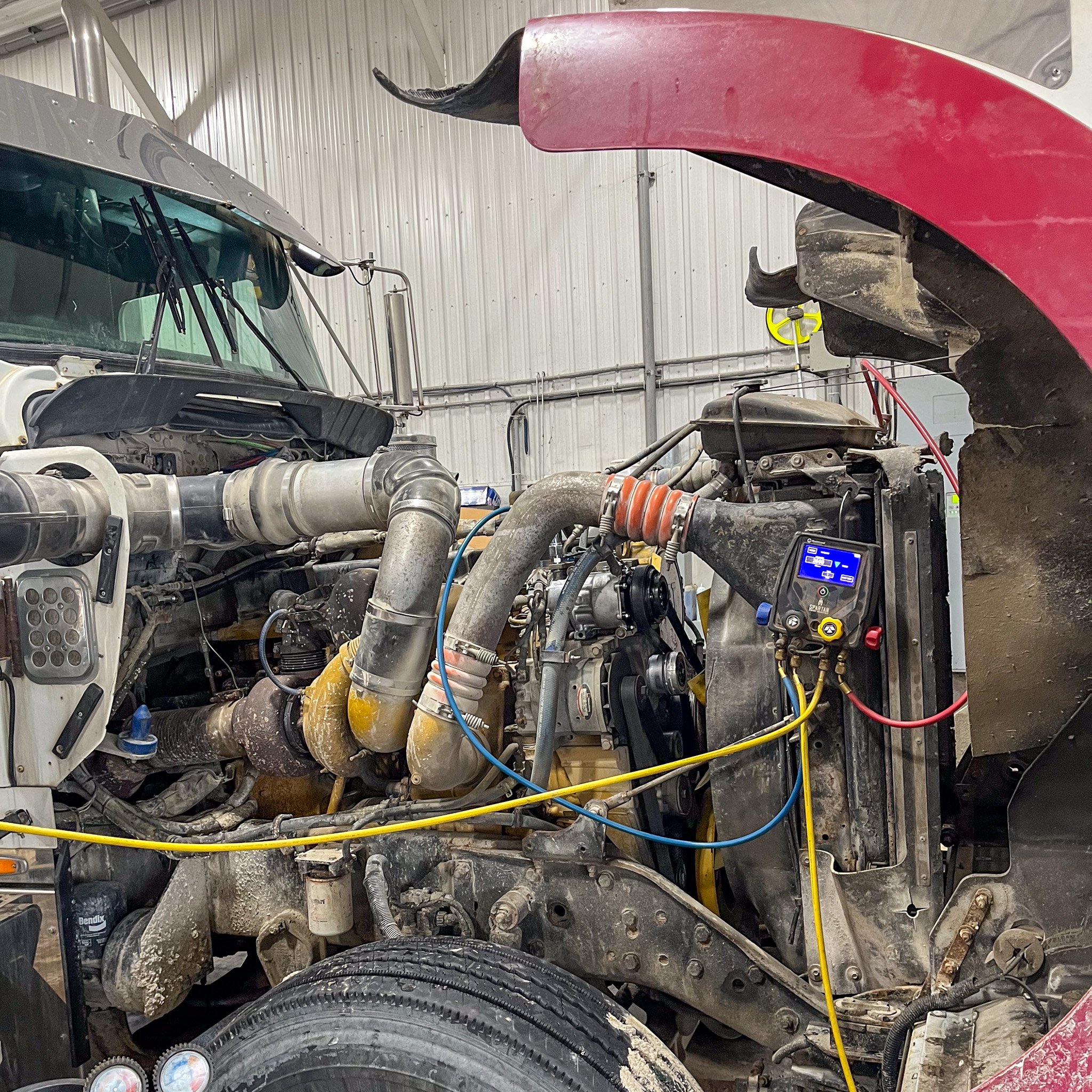 An industrial vehicle with its front panel removed, exposing engine components, pipes, wiring, and a digital display in a workshop.