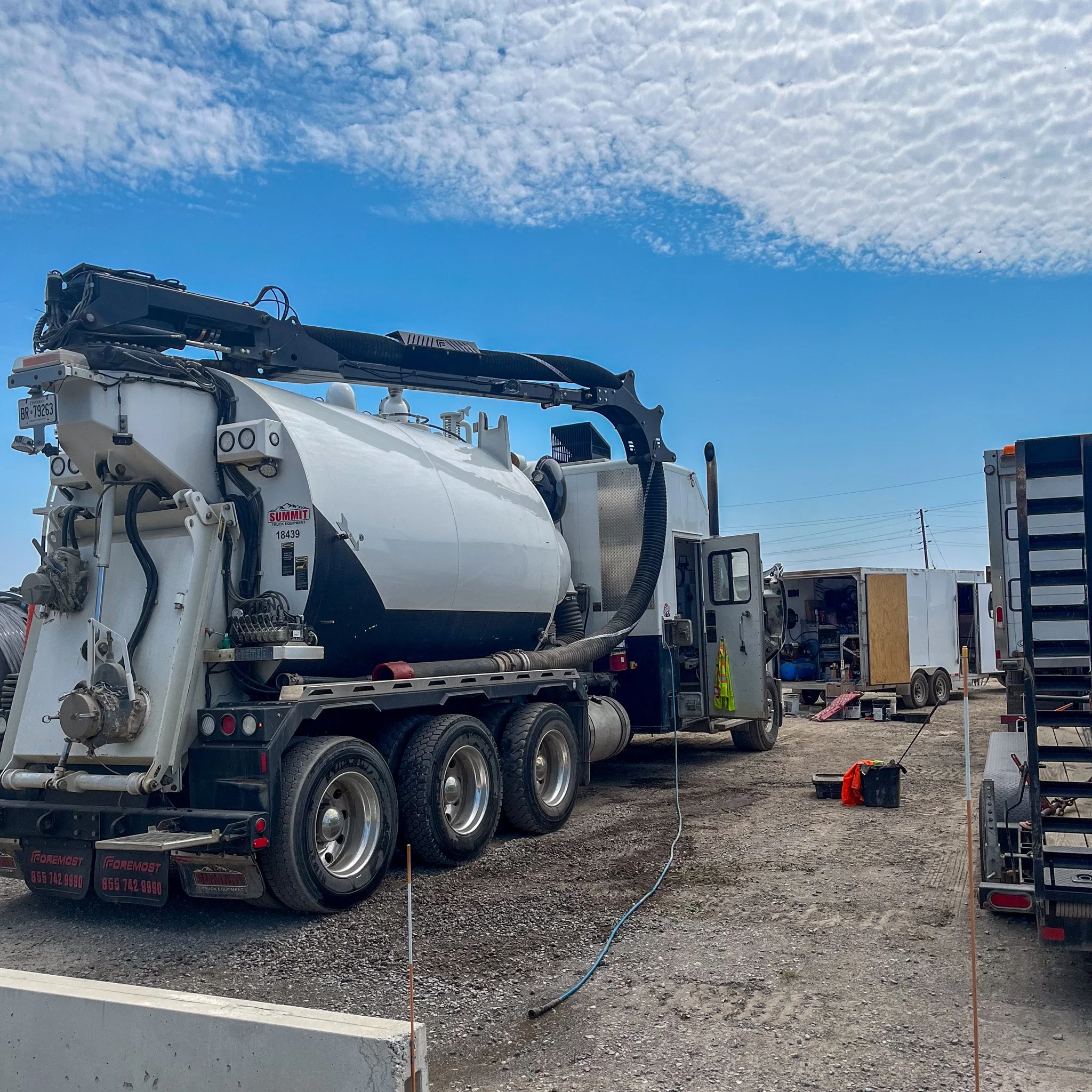A large white vacuum truck with a hose attached, parked at a construction site with other trucks and equipment nearby under a partly cloudy sky.