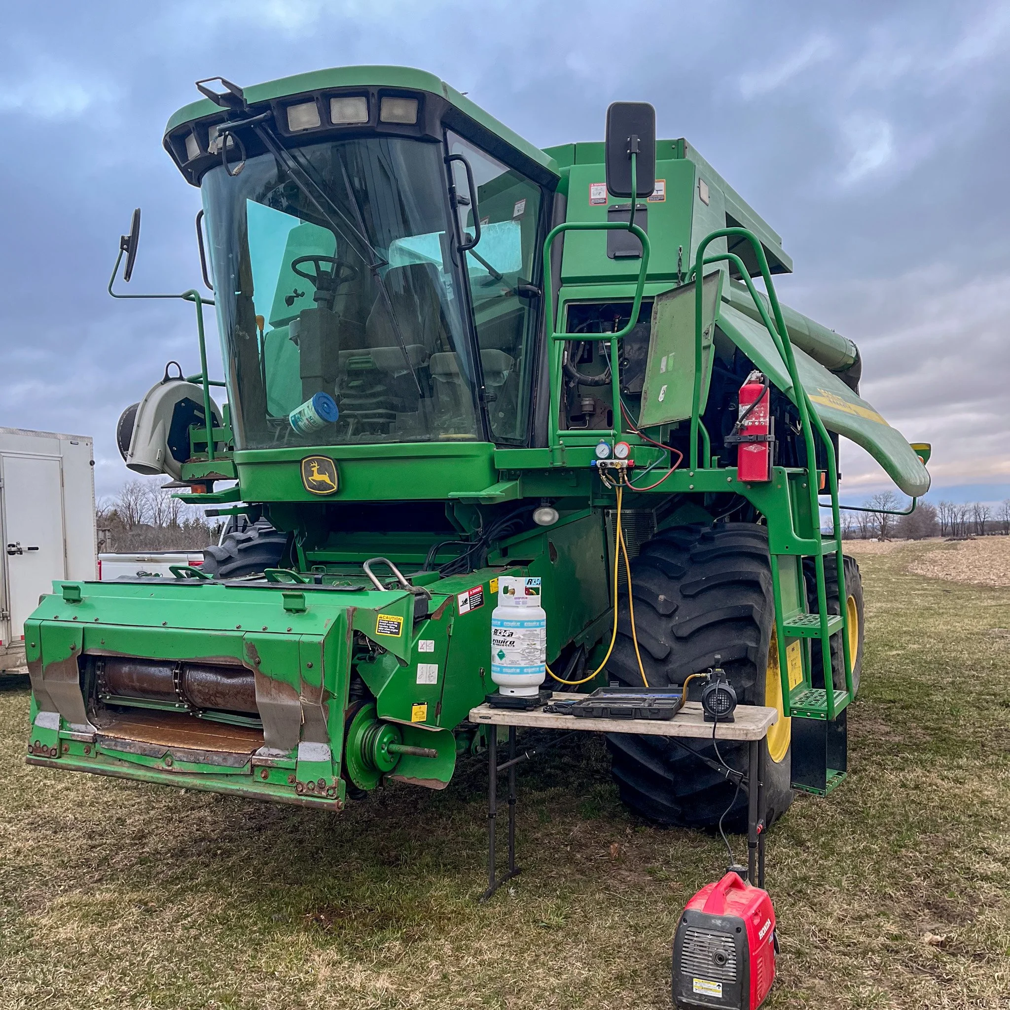 Large green John Deere combine harvester on grassy field with equipment and tools attached and nearby, under a cloudy sky.