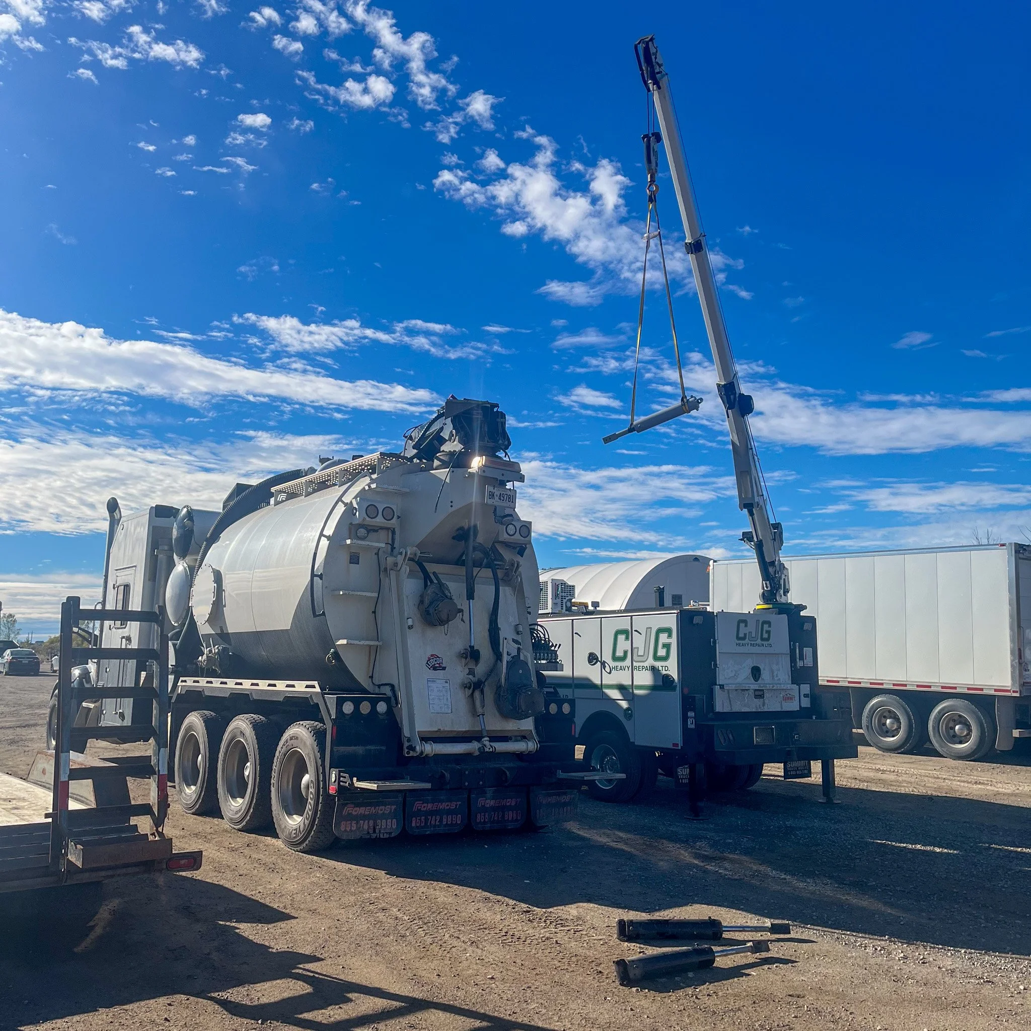 A large vacuum truck with a boom lift and piping on a dirt lot under a blue sky with scattered clouds, surrounded by other trucks.