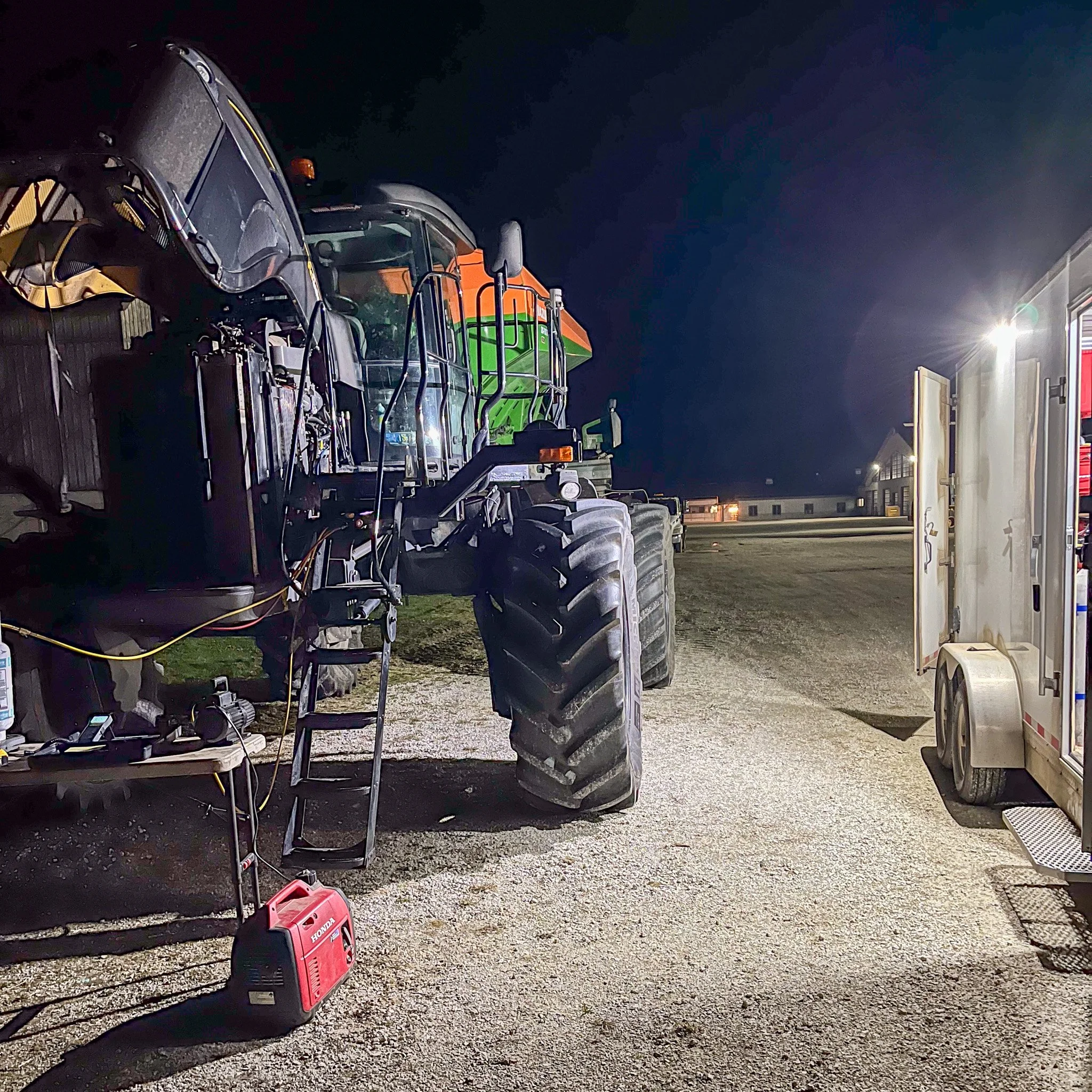 Nighttime scene showing a large orange and green agricultural combine harvesters parked on a gravel lot, with a bright light illuminating the area and a small white utility trailer on the right.
