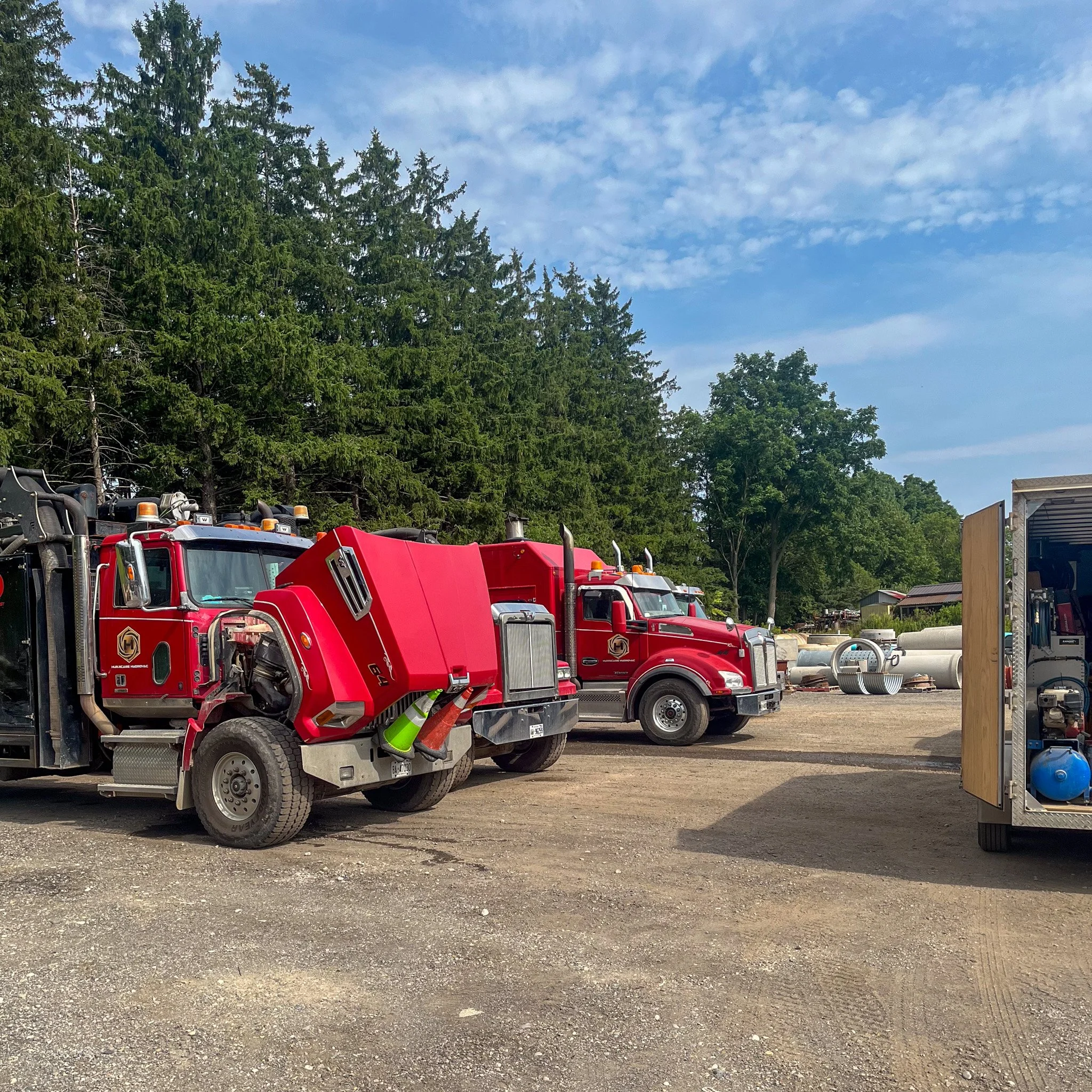 A row of red fire trucks parked on a dirt lot with trees and blue sky in the background.