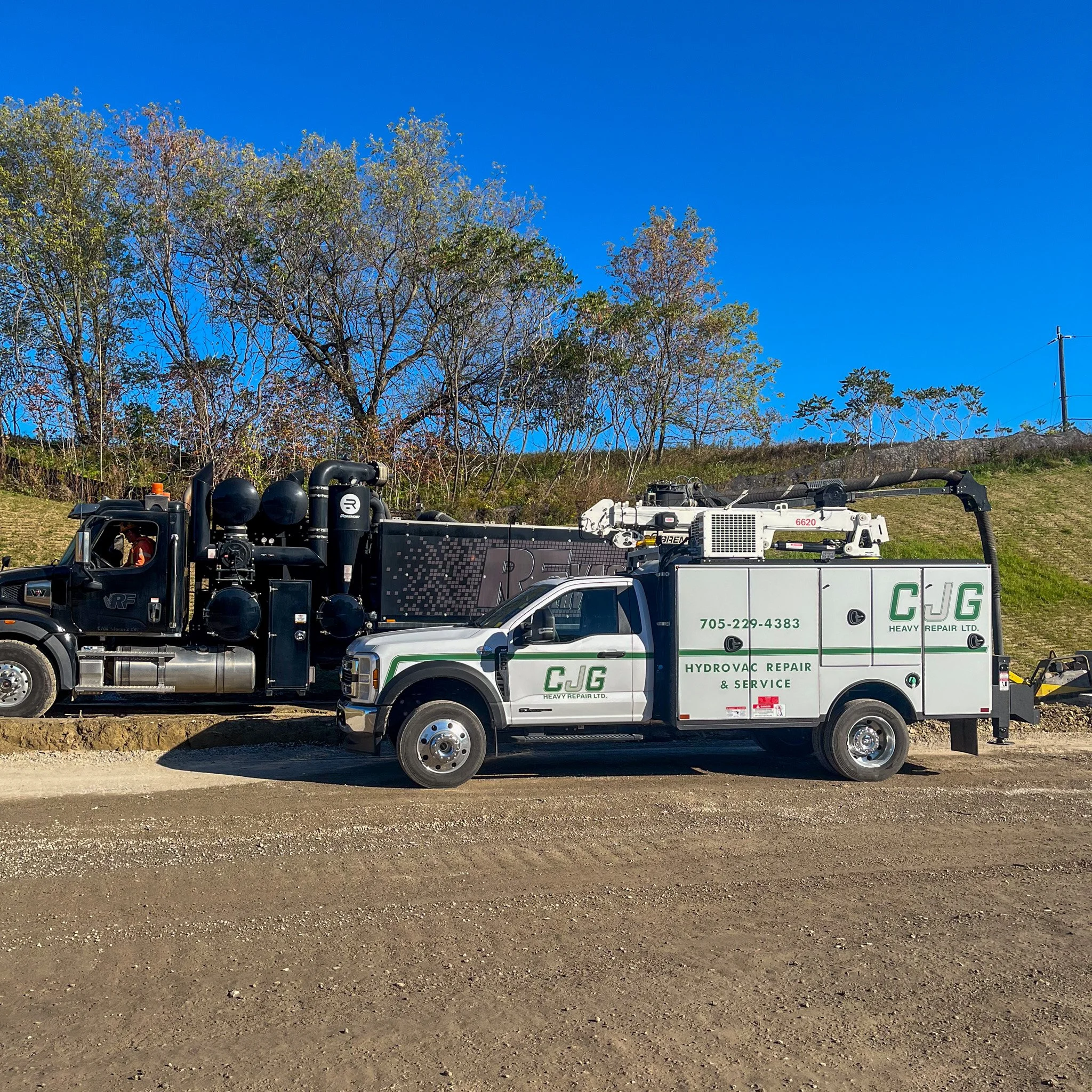 A hydro vacuum repair and service truck parked on a dirt road, with a large black industrial truck in the background and trees with fall foliage under a clear blue sky.