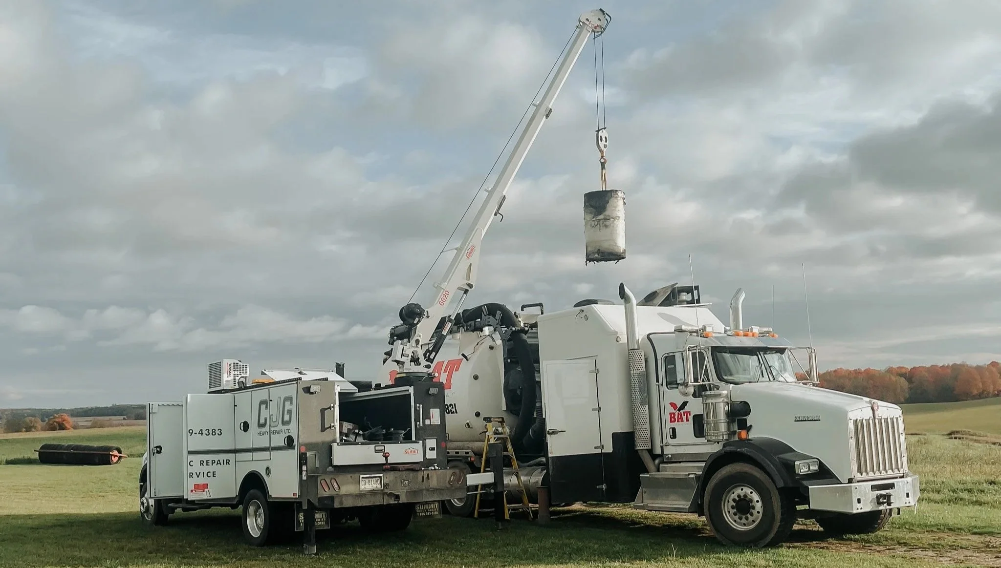A utility truck with a crane lifting a large, weathered barrel in an open field under a cloudy sky.