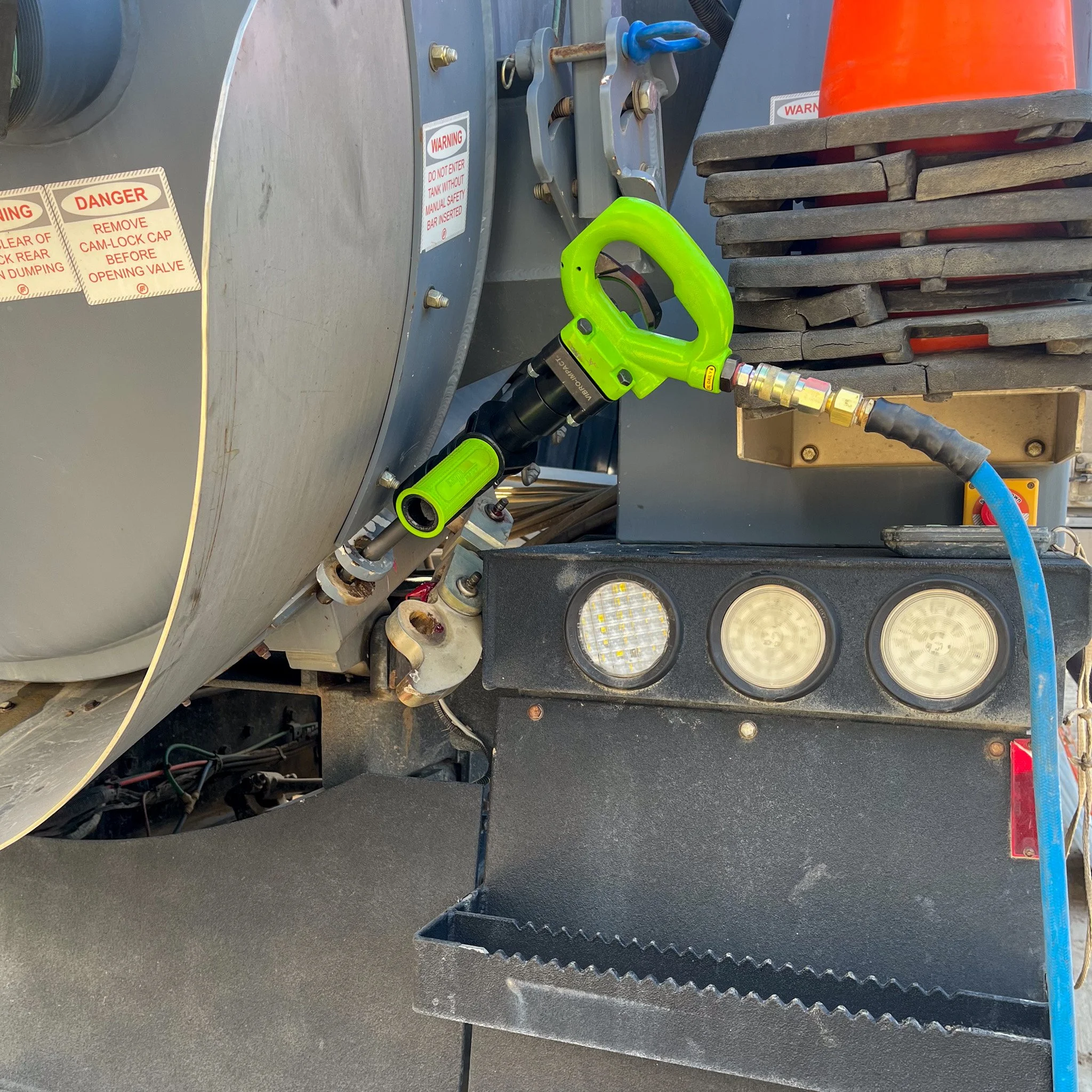 Close-up of a truck's front bumper with three round lights, a green hydraulic or pneumatic tool attached to a metal component, and a blue hose connected to a fitting. Stacked pallets and safety warning labels are visible in the background.