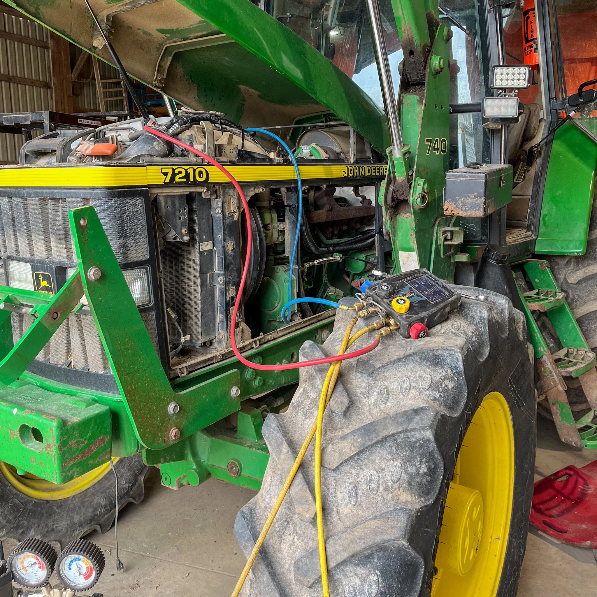 A green John Deere tractor with its engine cover open, exposing the engine components, and a diagnostic tool connected to the engine with wires. The tractor has yellow wheels and appears to be in a garage or workshop setting.