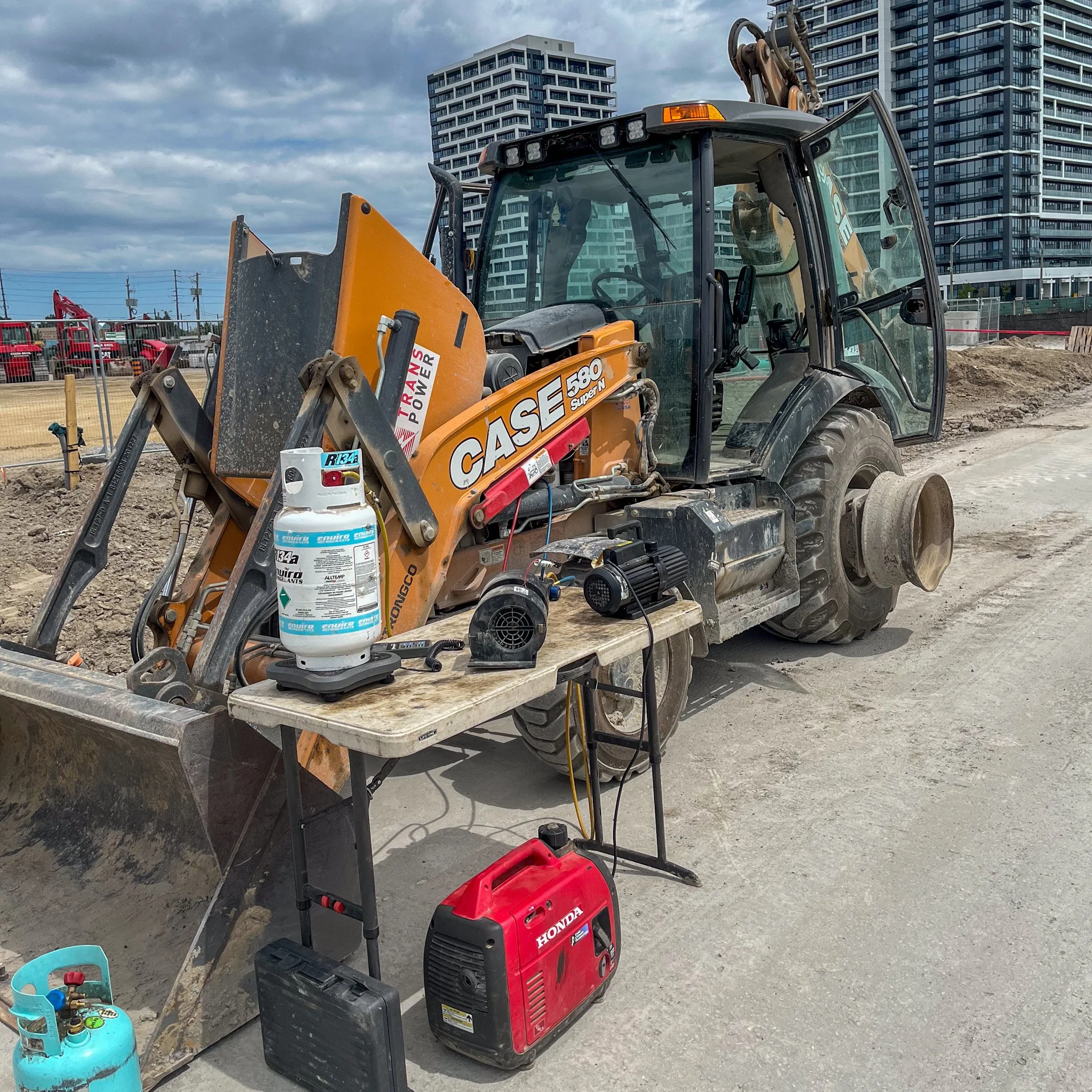 Construction site with a Case backhoe loader, a table with a propane tank, a red Honda generator, and various tools and equipment, with buildings and a cloudy sky in the background.