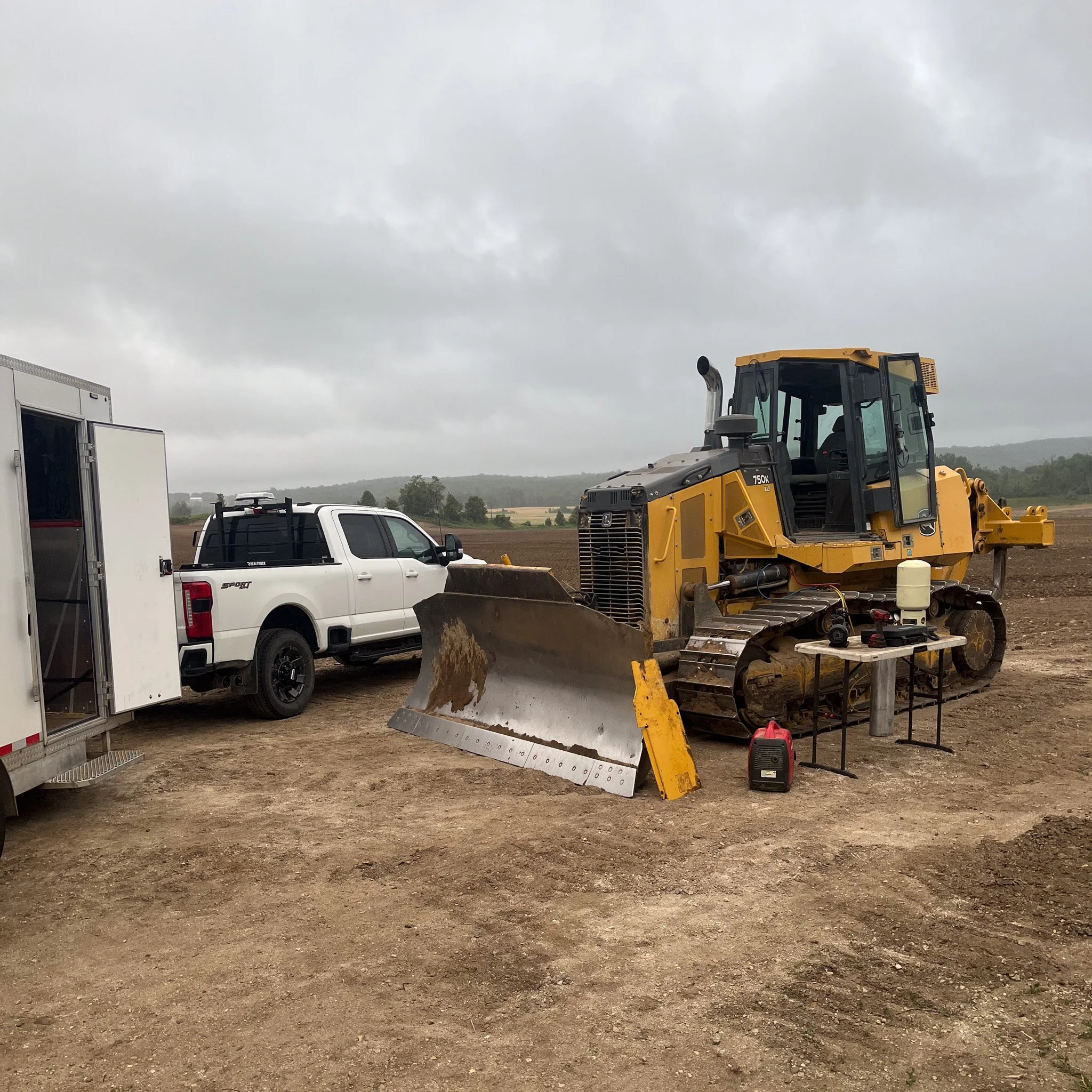 A yellow bulldozer on a dirt field with a white pickup truck and a white trailer nearby, under cloudy skies.
