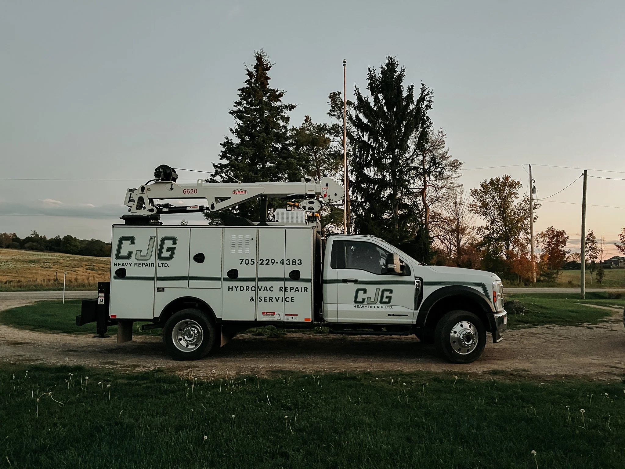 A white service truck labeled 'CJG Heavy Repair Ltd.' and 'Hydrovac Repair & Service' parked on a dirt patch near grassy fields and trees, with utility poles and wires in the background during dusk or dawn.