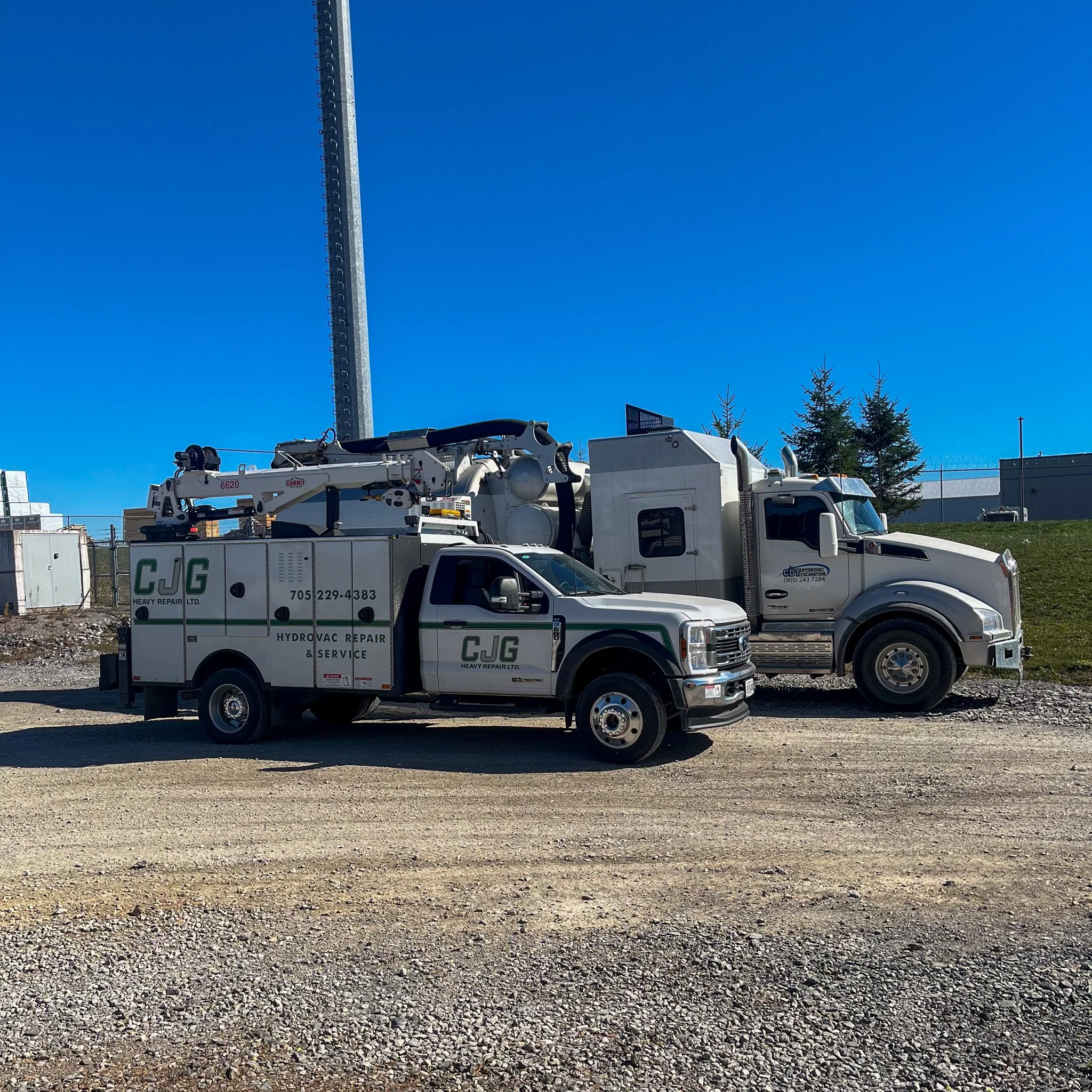 Two service trucks parked on a gravel lot, with the sky and some trees in the background. One truck is a white utility truck labeled 'CJG Heavy Repair Ltd.' and the other is a larger white truck with equipment on top.