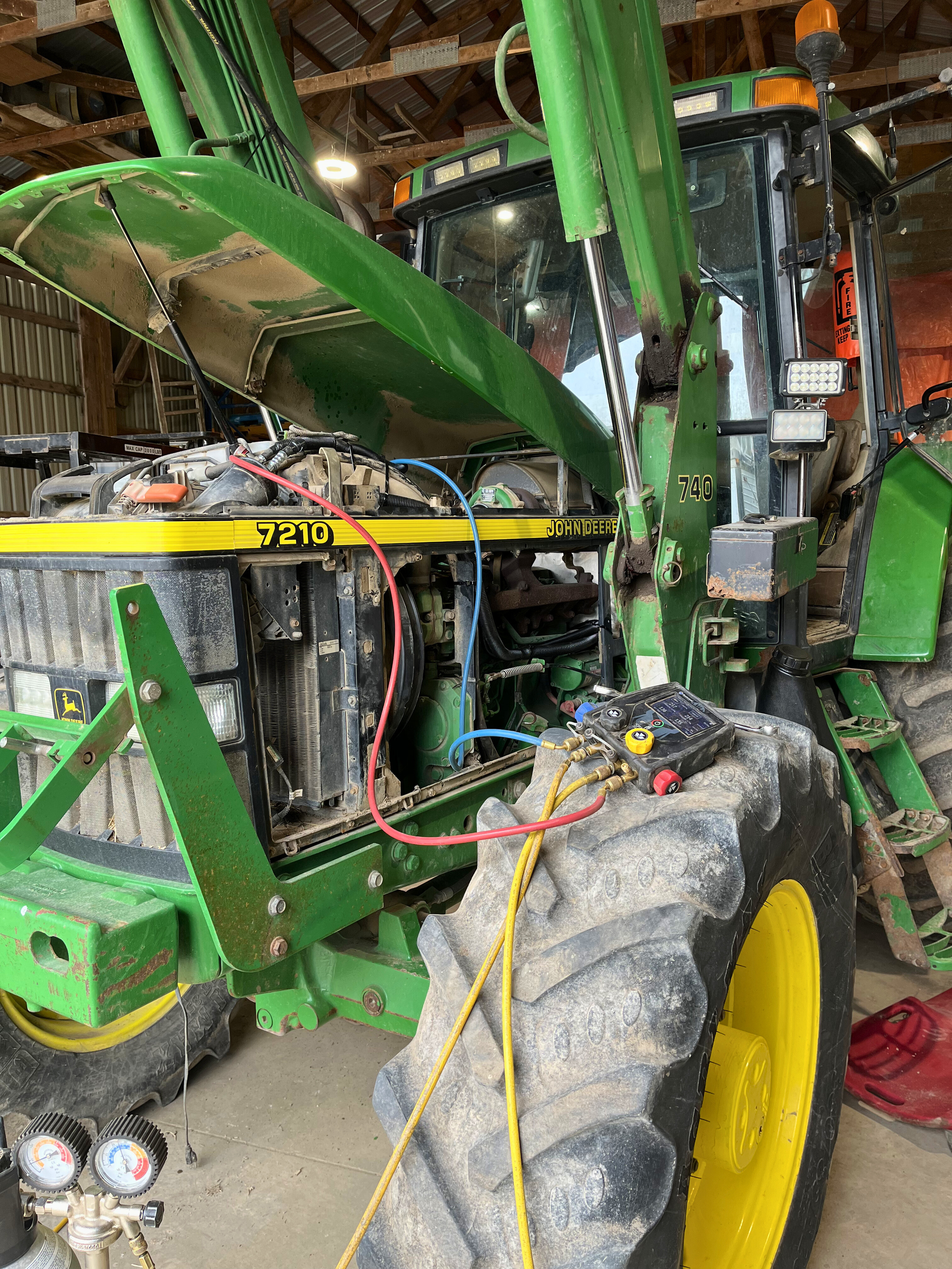 A green John Deere tractor with its hood partially open, showing the engine. The tractor is in a barn or shed with wooden beams overhead, and some tools and equipment are visible.