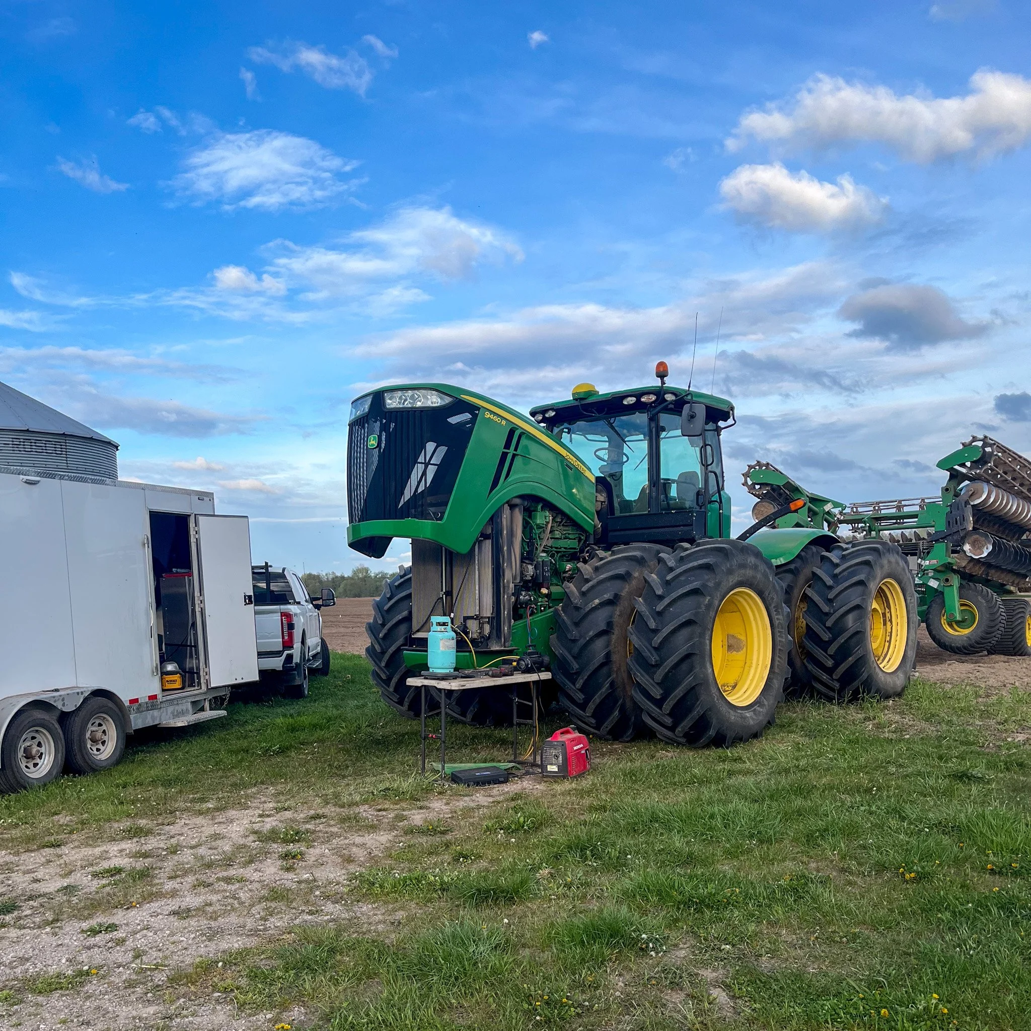 Green John Deere tractor with large wheels and attached equipment, parked on grass next to a trailer and a white pickup truck under a partly cloudy sky.