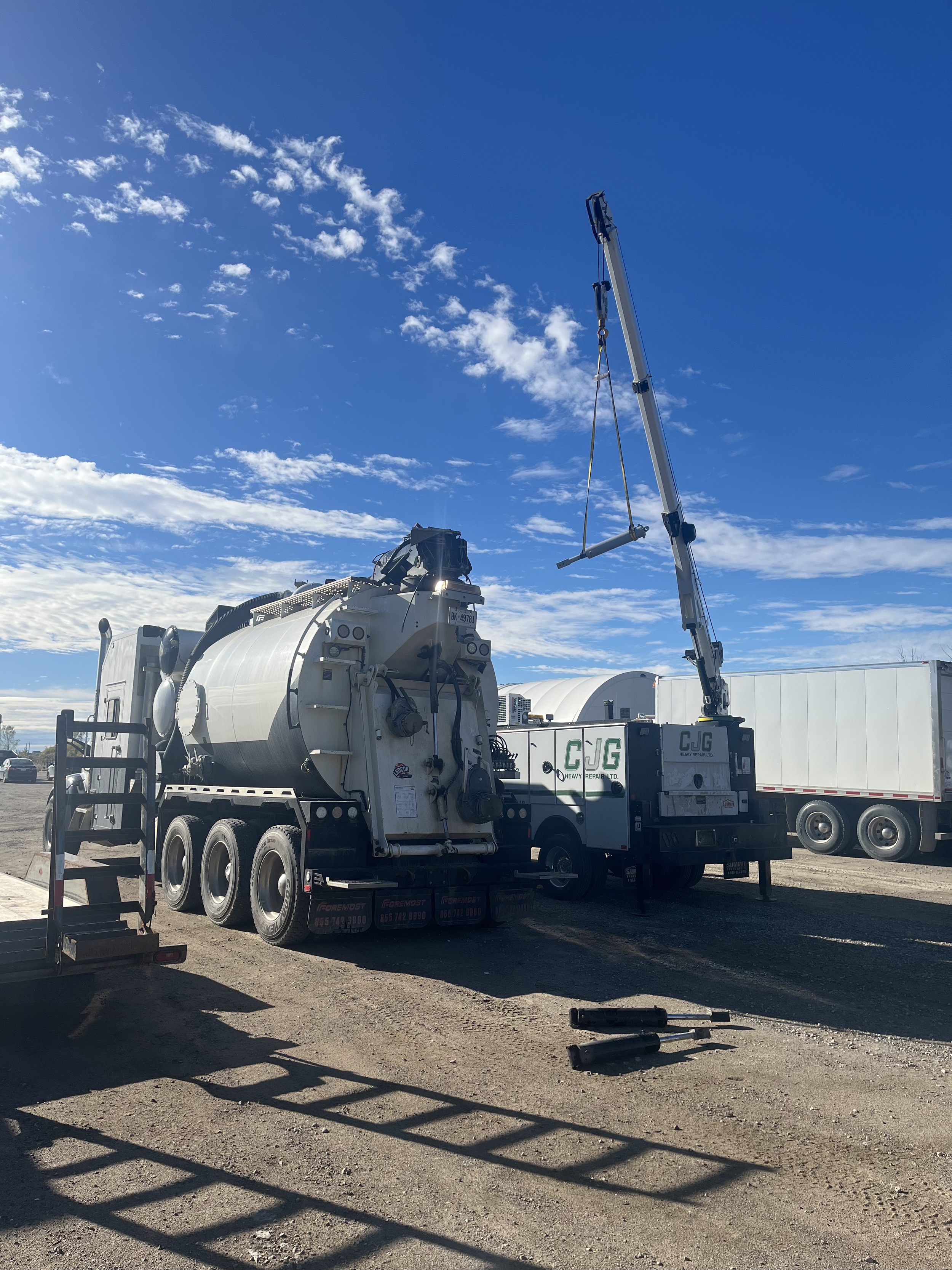 A heavy-duty vacuum truck with a large cylindrical tank is parked on a construction site under a blue sky with clouds. The truck has a crane extended and a ladder on the side, with a few tools on the ground nearby.