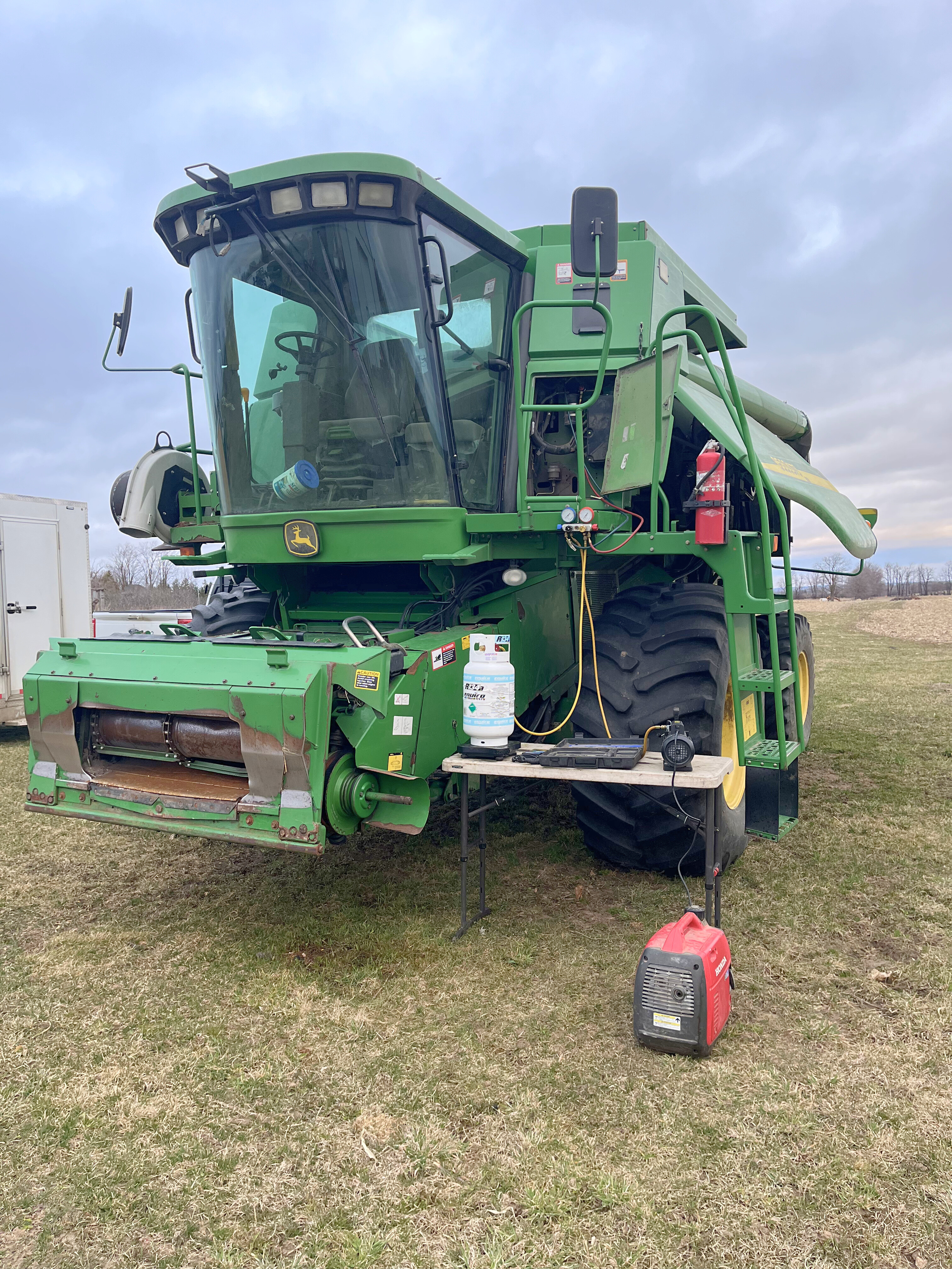 a large green John Deere combine harvester parked on grass with maintenance equipment and a propane tank nearby
