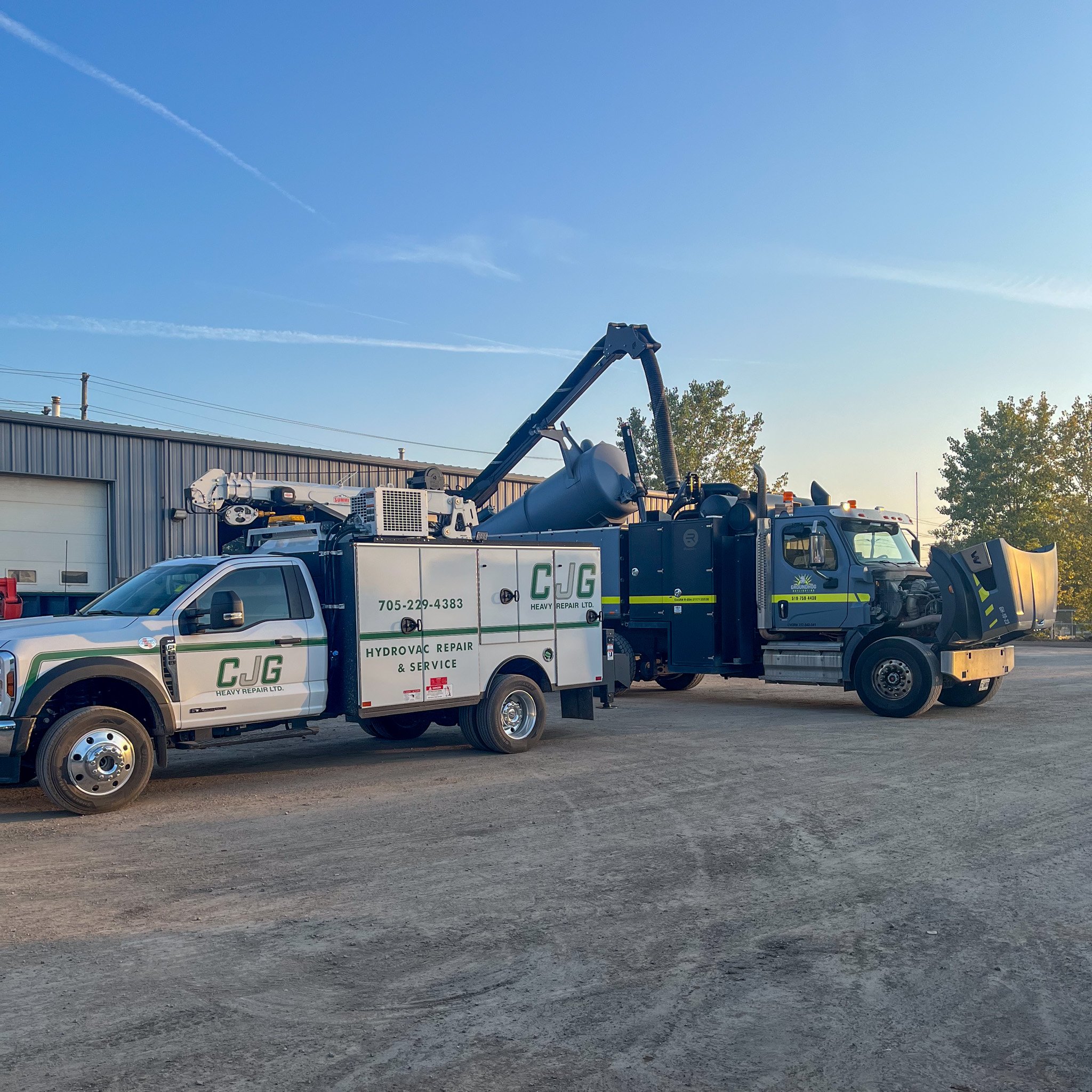 A large black tow truck with a hydraulic lift and tools parked next to a service van with CJG Heavy Repair logos and contact information, outdoors on a dirt lot with a warehouse and trees in the background.