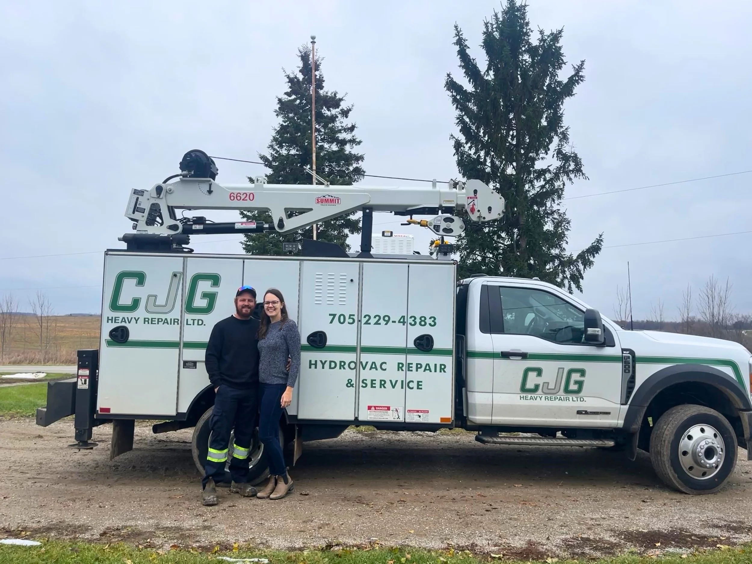 Two people standing in front of a white service truck labeled CJG Heavy Repair Ltd. with a hydraulic lift on top. The individuals are smiling, the man is wearing a black jacket and work pants, and the woman is wearing a gray sweater and jeans. The background shows overcast sky, trees, and an open field.