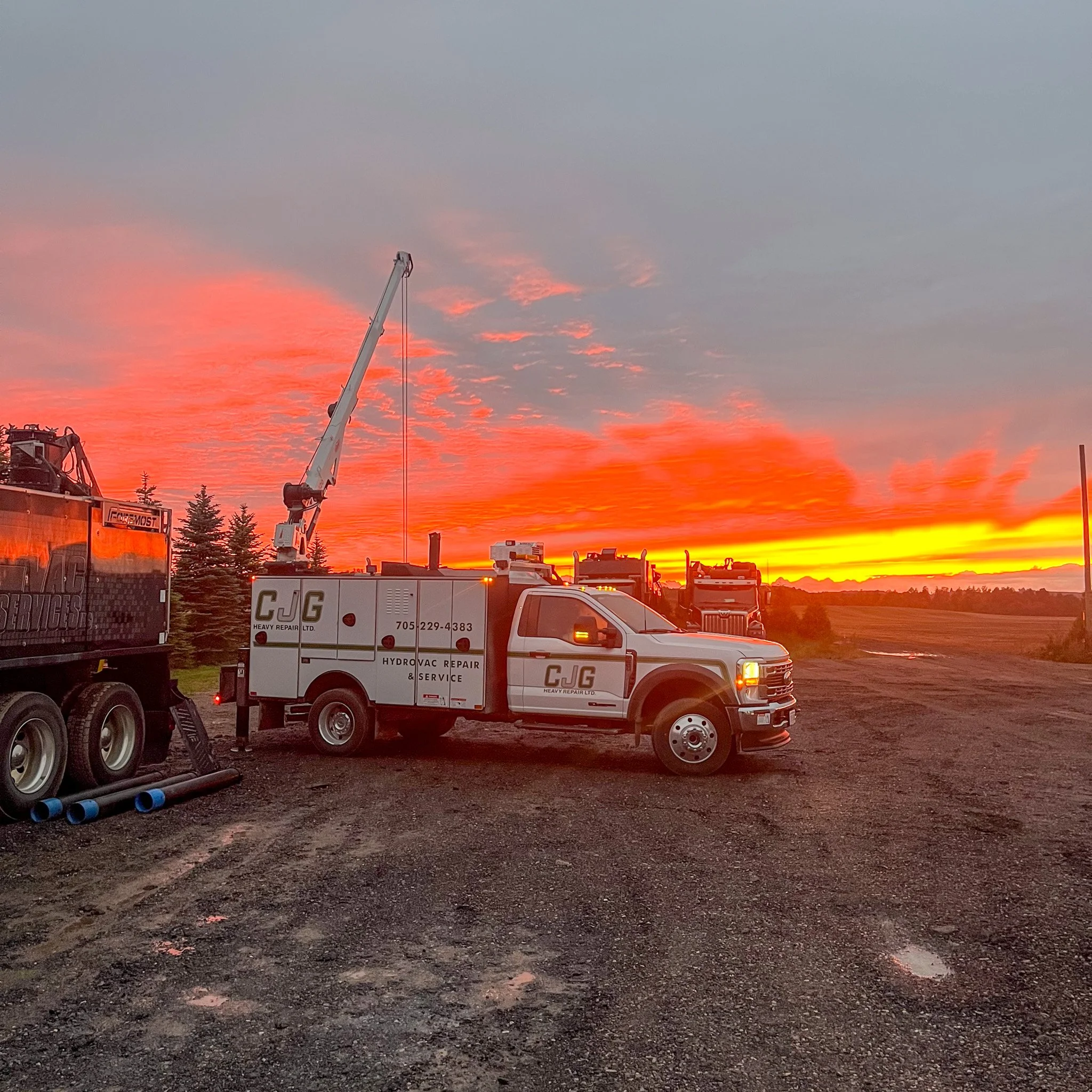 Several repair trucks and equipment parked on a gravel lot during sunset, with orange and yellow sky and a few trees in the background.