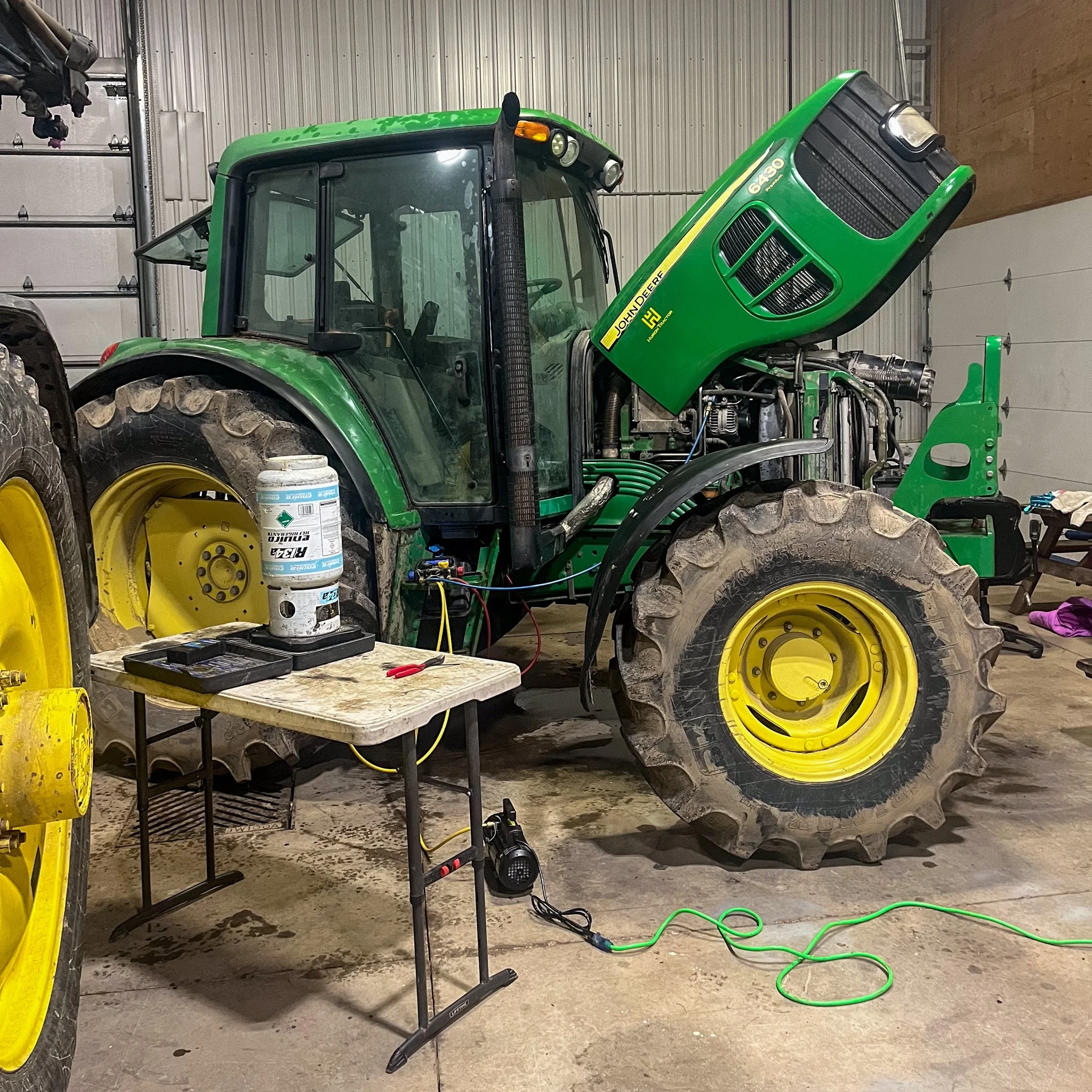 A green John Deere tractor inside a workshop with an open hood, connected to diagnostic equipment on a small table, with tools and cords on the floor.