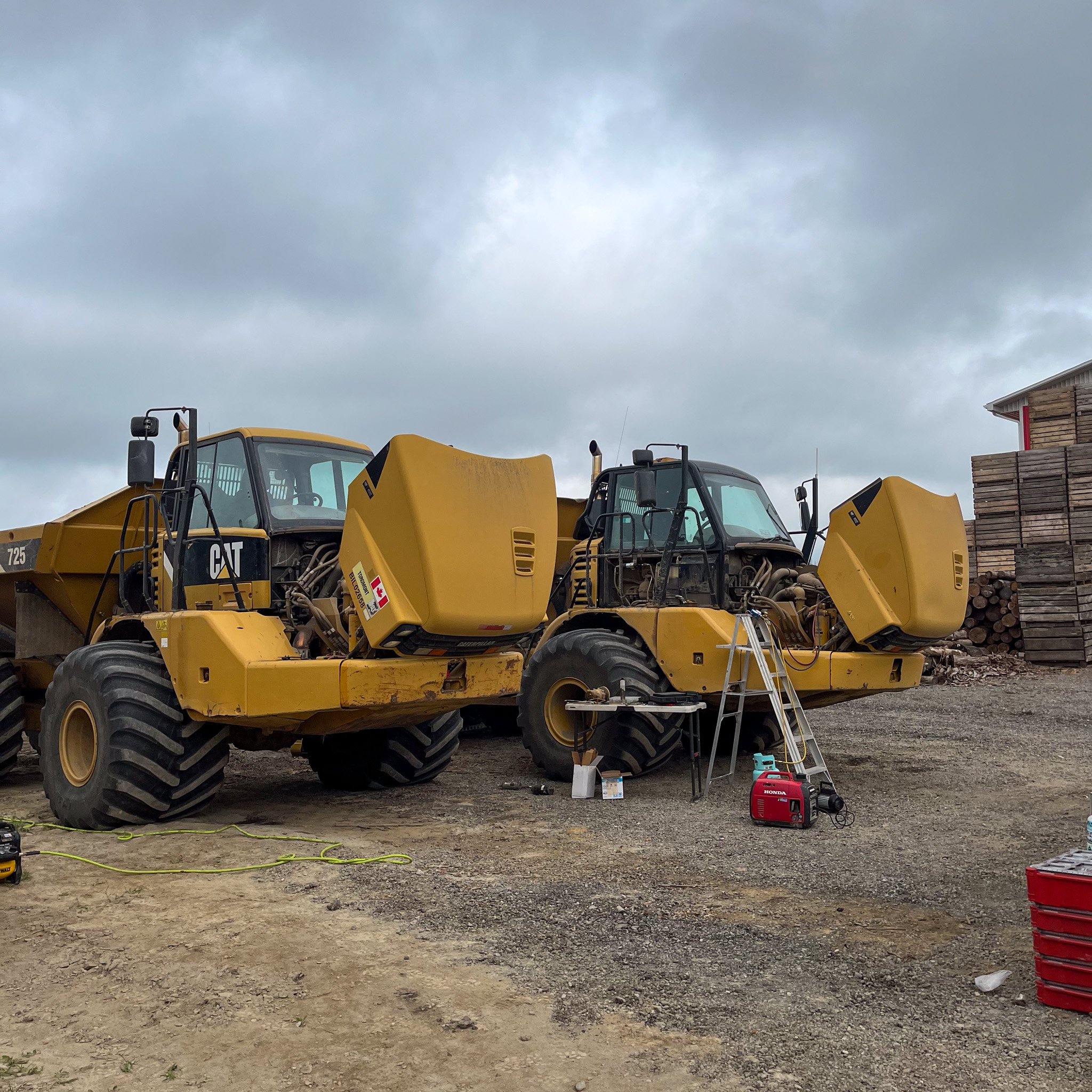 Two yellow CAT construction vehicles parked on a dirt ground with a ladder, toolbox, and construction equipment nearby, and stacked wood in the background under a cloudy sky.